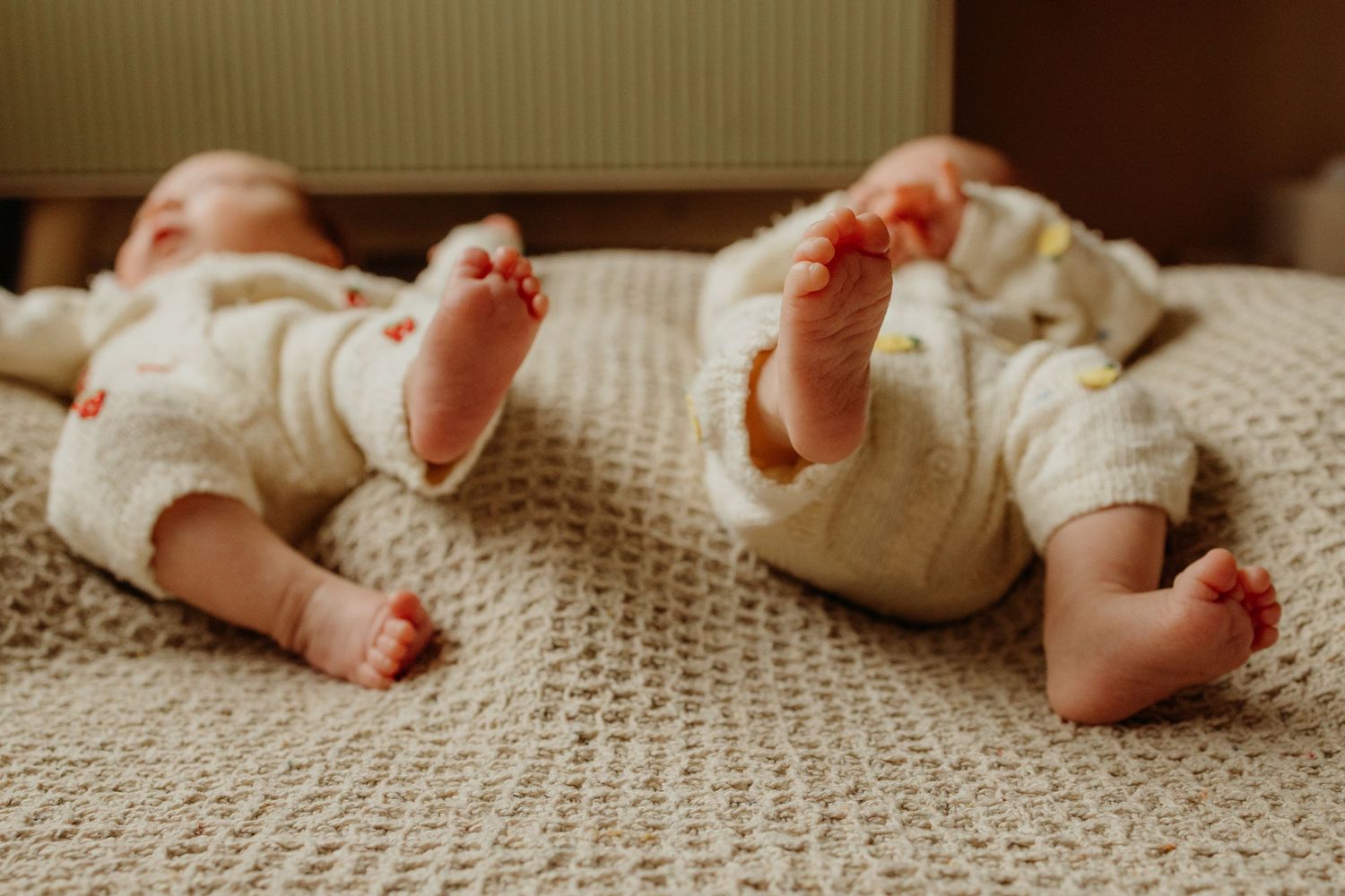 Two newborn twins lying side by side on a cream knit blanket.