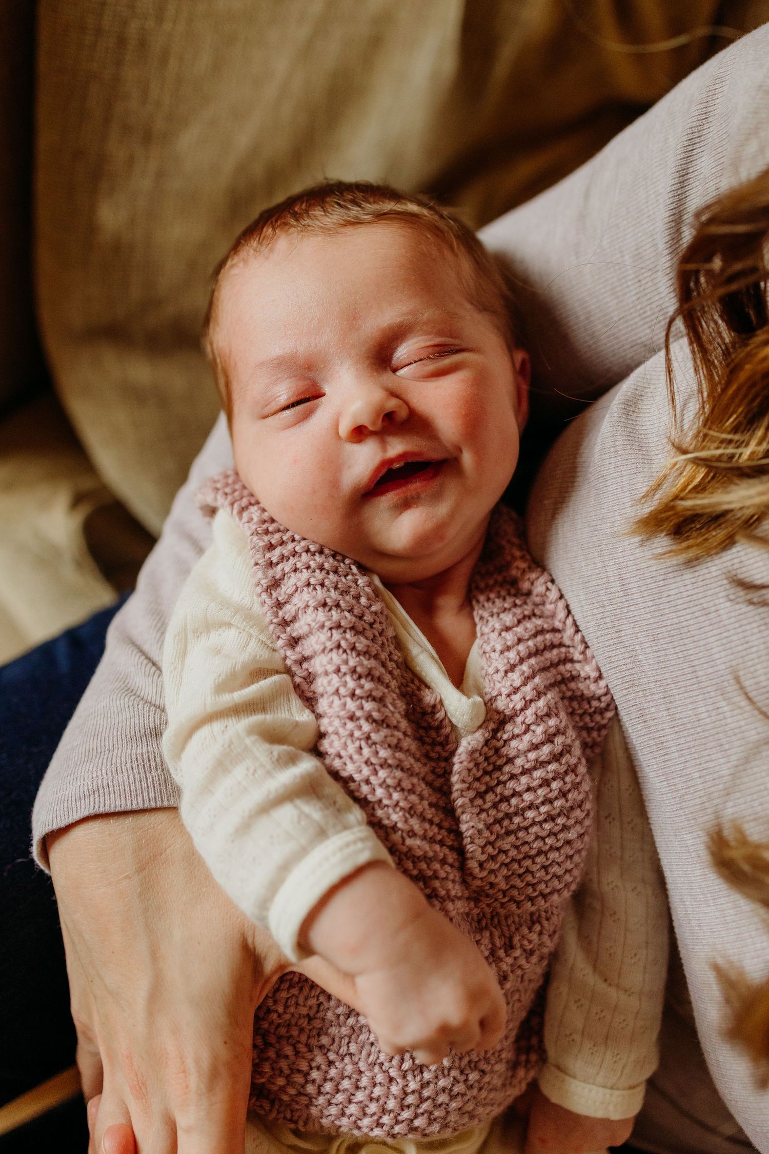 Smiling newborn baby in a pink knit wrap resting in a parent's arms.