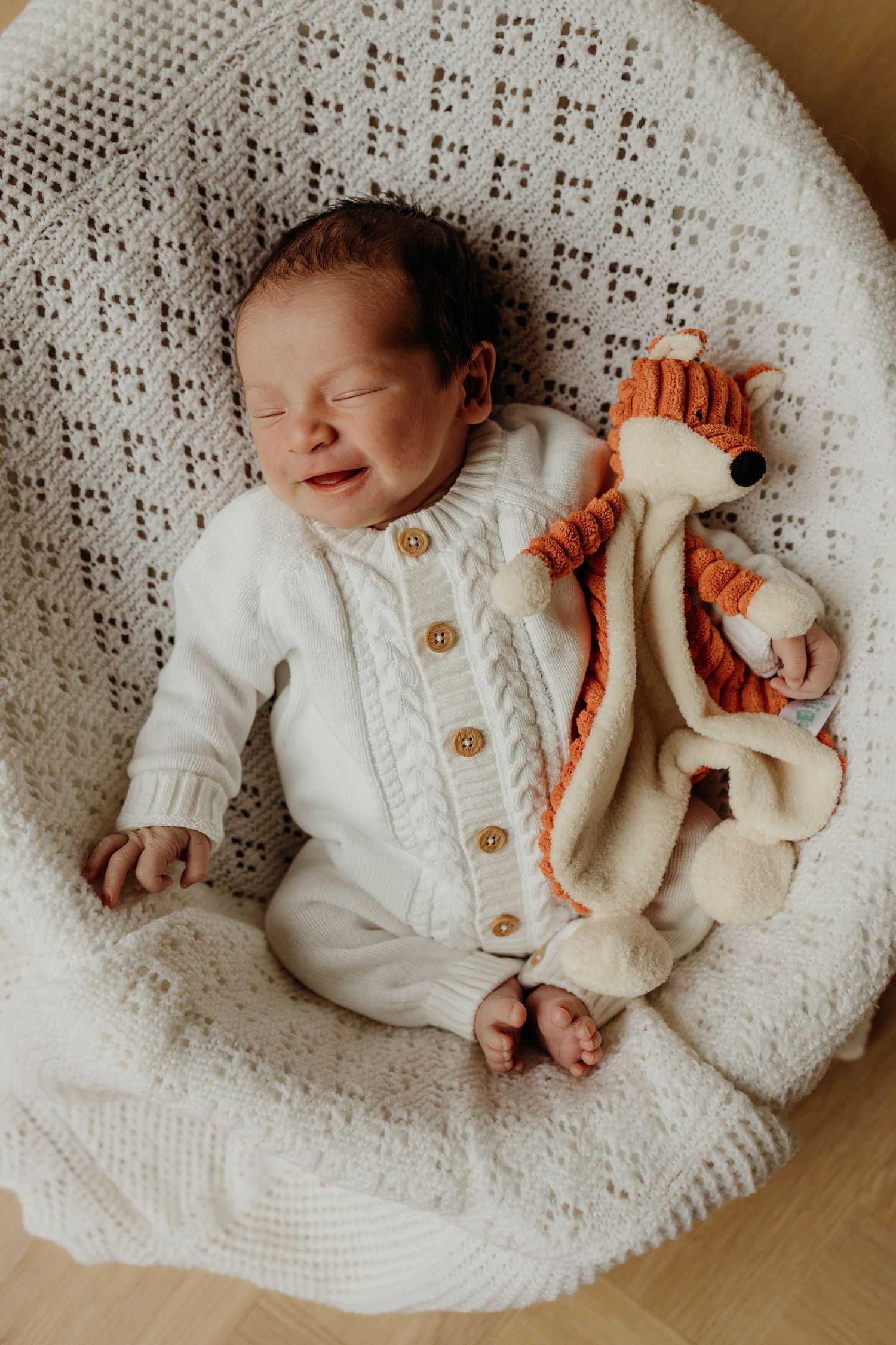 Sleeping newborn in white cable-knit outfit beside an orange stuffed animal in a basket.