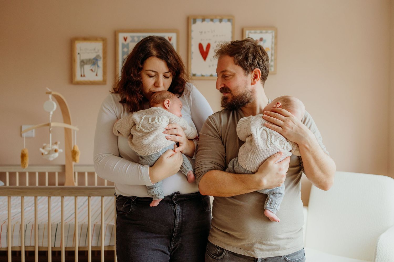 A couple stands in a nursery holding twin babies, surrounded by soft decor and a wooden crib.