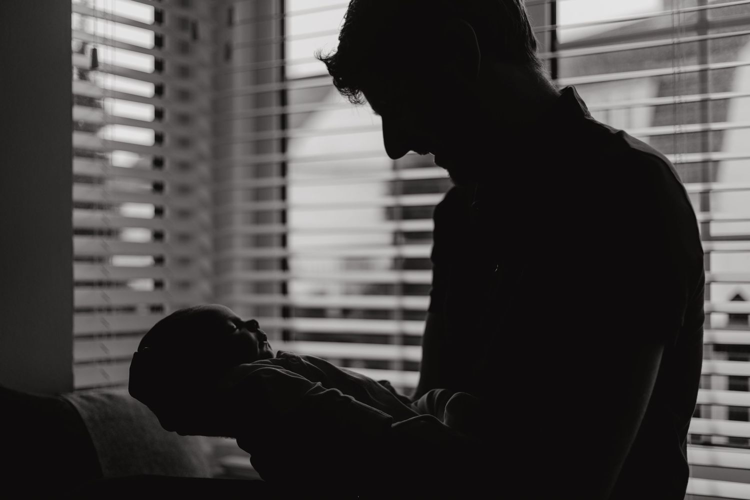 Black and white silhouette of a father holding his newborn baby near a window with blinds.