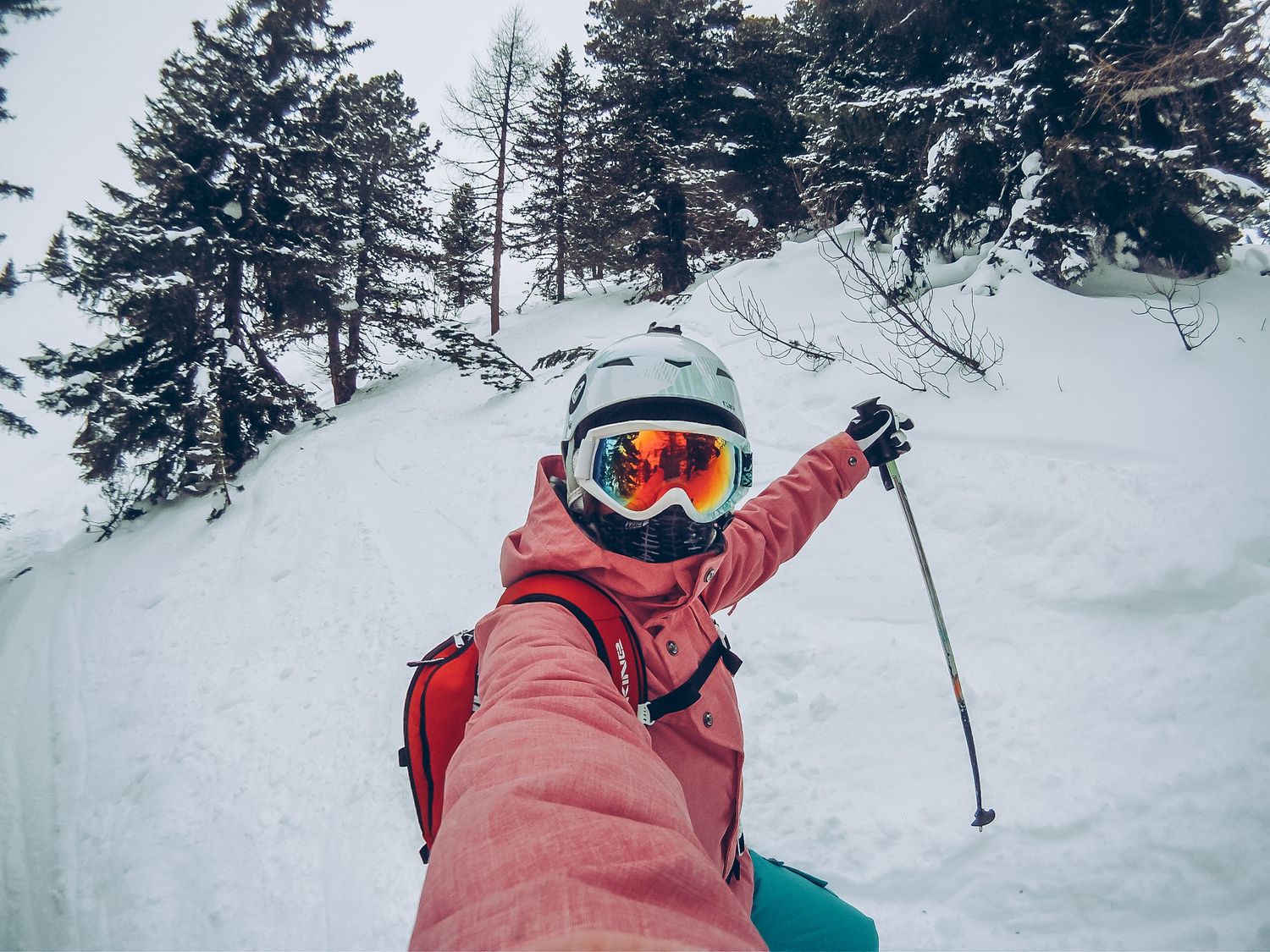 A skier in a pink jacket and helmet takes a selfie on a snowy mountain slope surrounded by pine trees.
