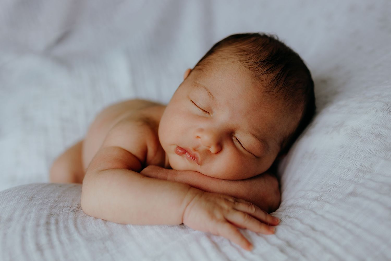 A peaceful newborn baby sleeps with arms folded beneath its chin on a soft white blanket.