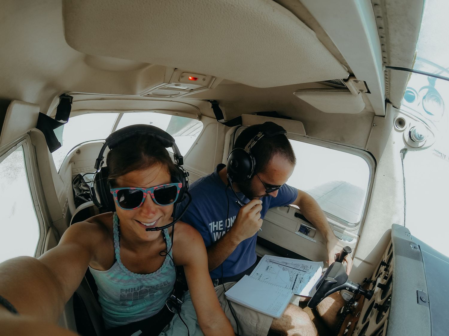 Two pilots wearing headsets take a selfie inside a small aircraft cockpit during a sunny flight.