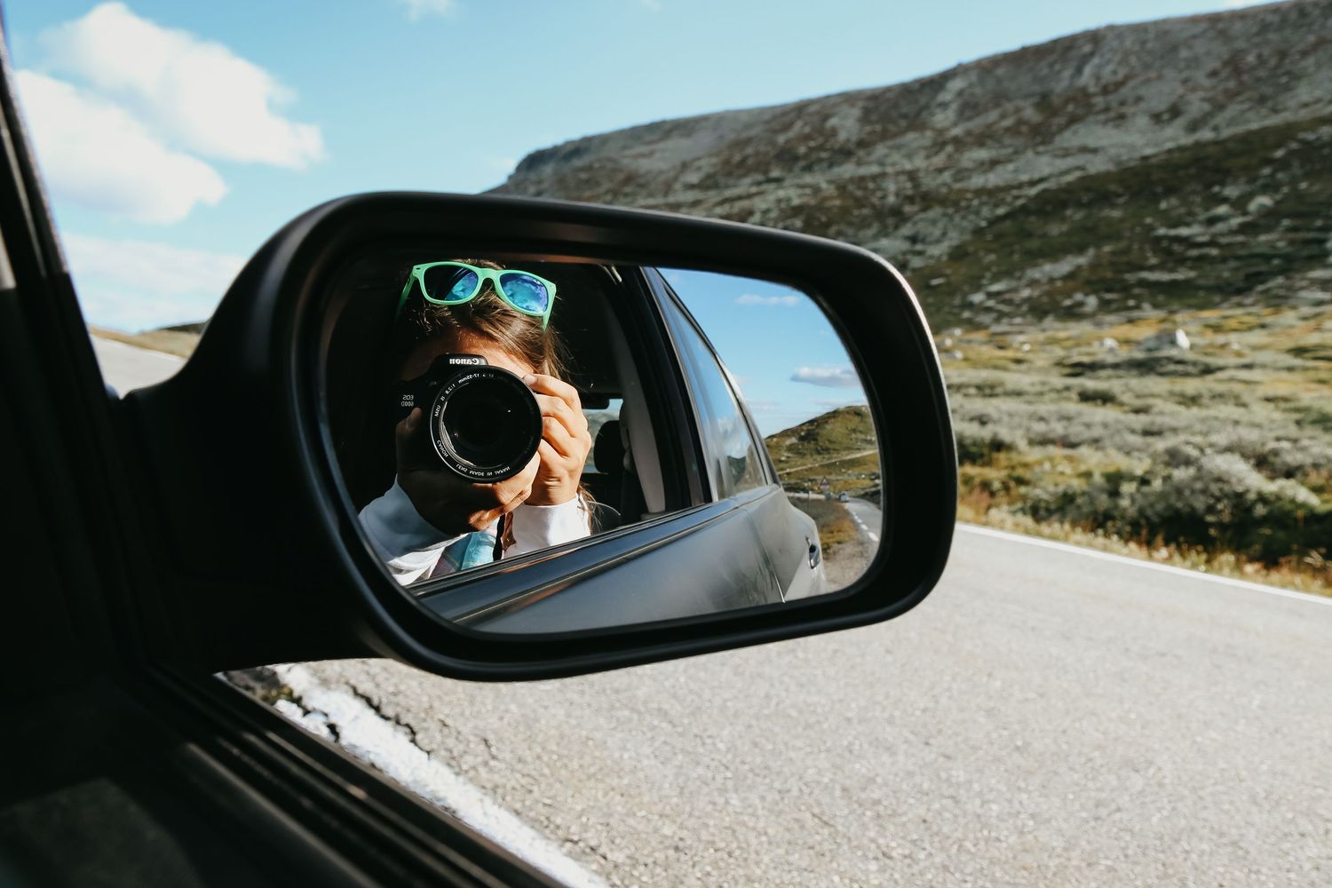 A woman with colorful sunglasses photographs herself in a car side mirror on a scenic mountain road.
