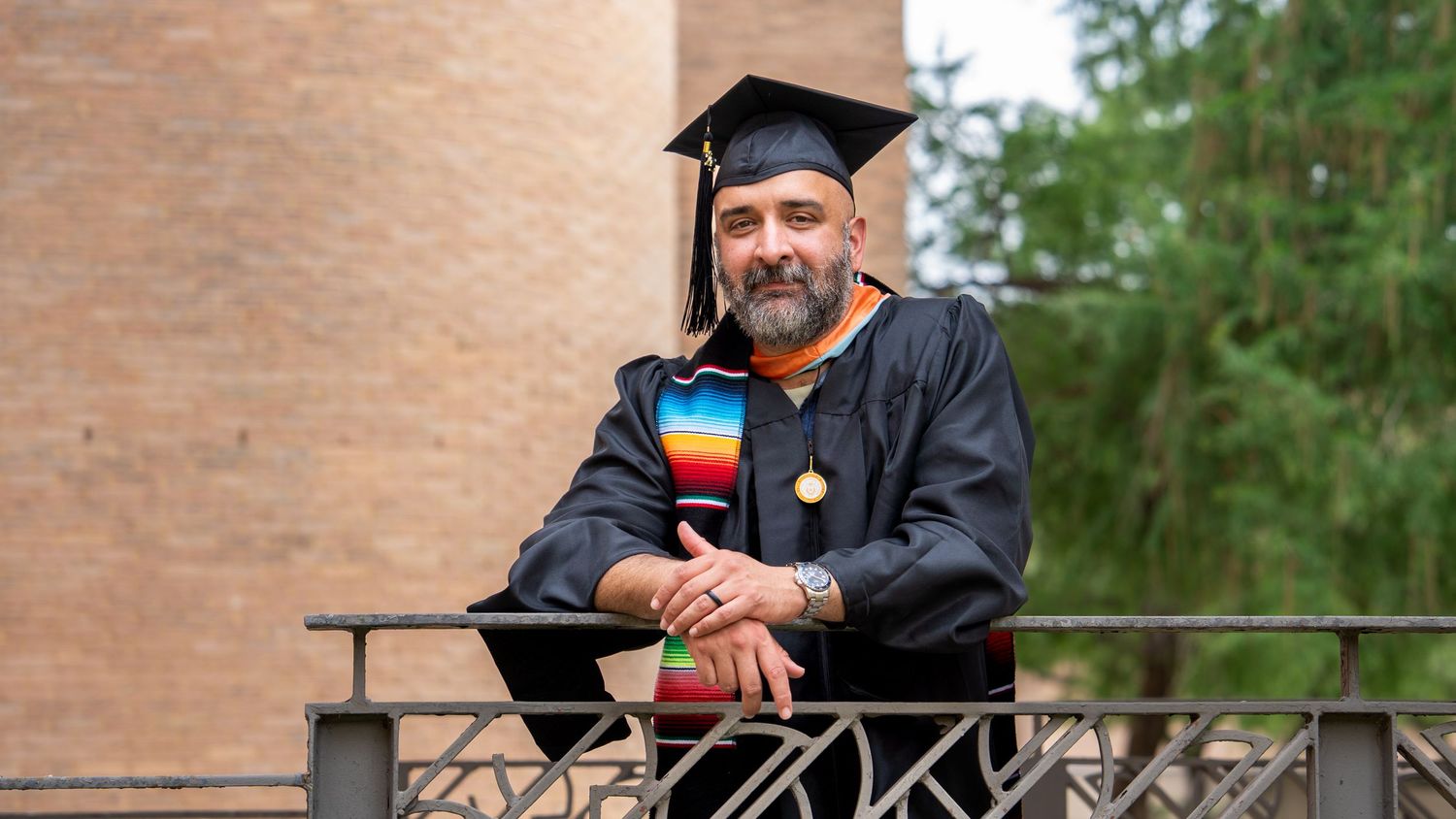 Man in graduation cap and gown with rainbow stole leaning on railing at outdoor university commencement ceremony.