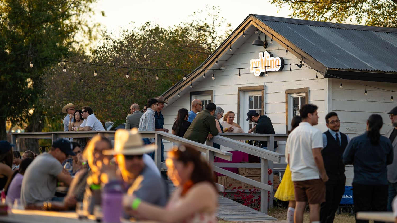 Crowd of people socializing on outdoor deck and patio of small white building at golden hour sunset.