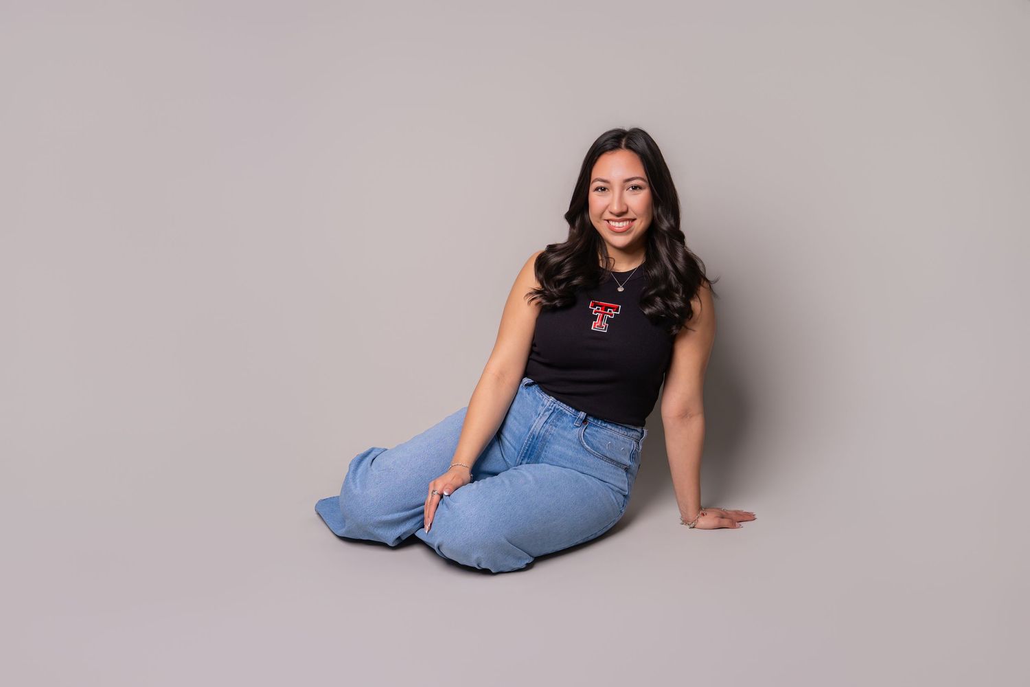Young woman with long dark hair sitting on floor wearing black Texas Tech tank top and jeans smiling.