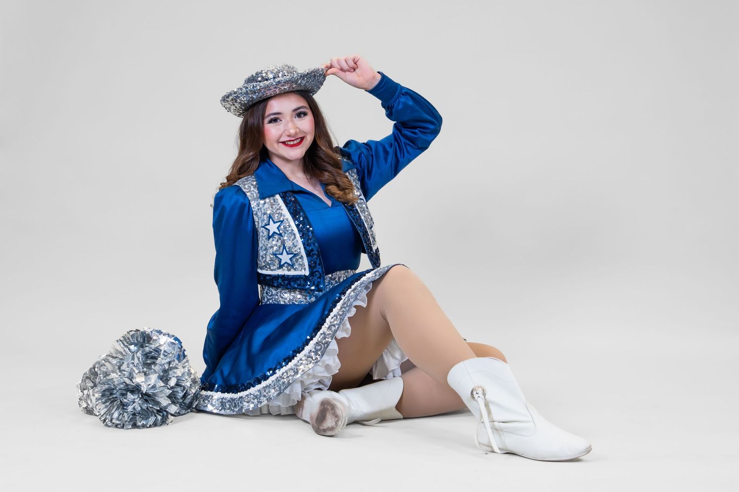 Young woman in blue silver majorette uniform and cowboy hat sitting with pom poms on white background.