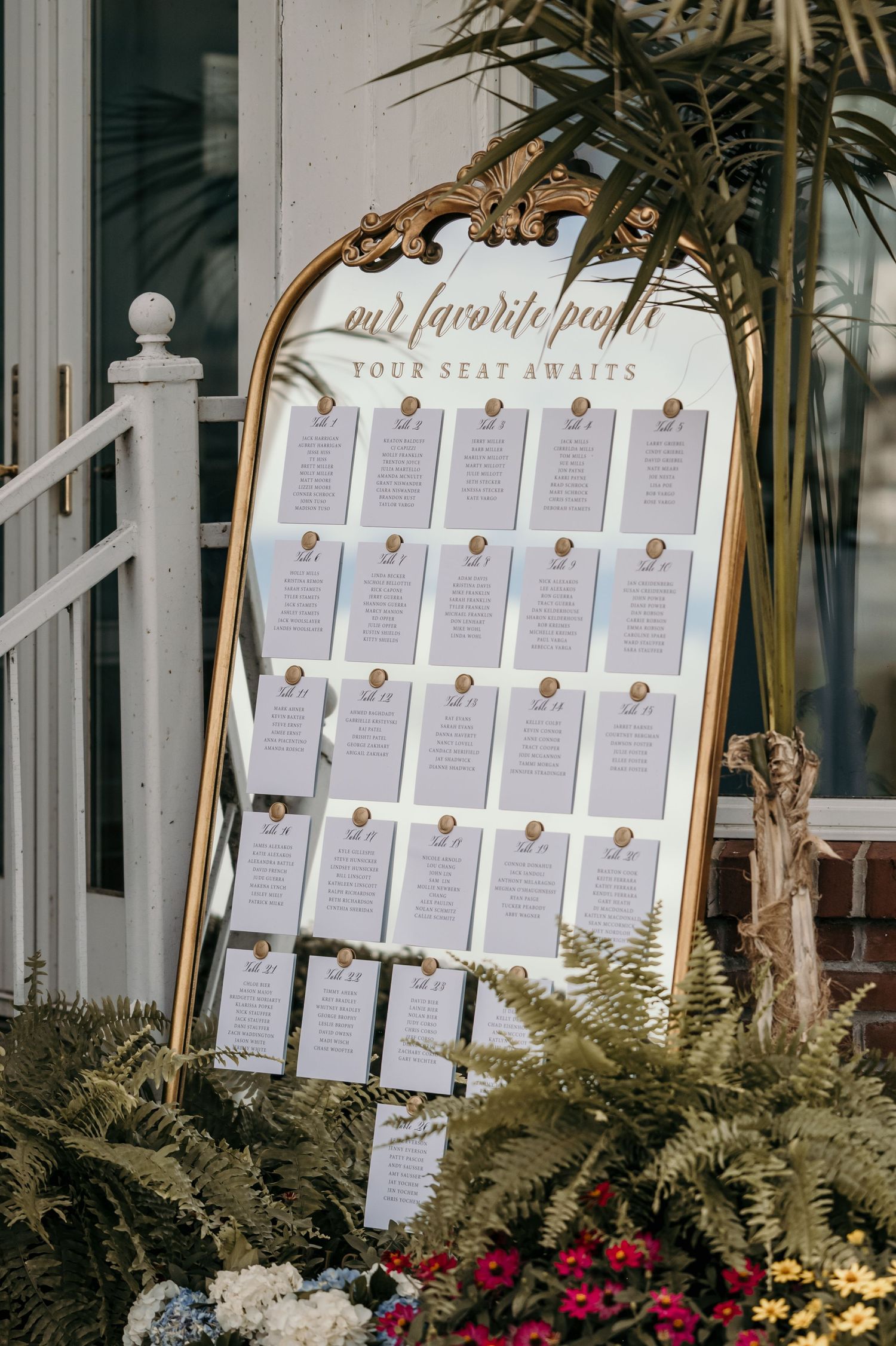 Elegant gold ornate mirror seating chart display with white cards reading our favorite people your seat awaits at outdoor wedding.