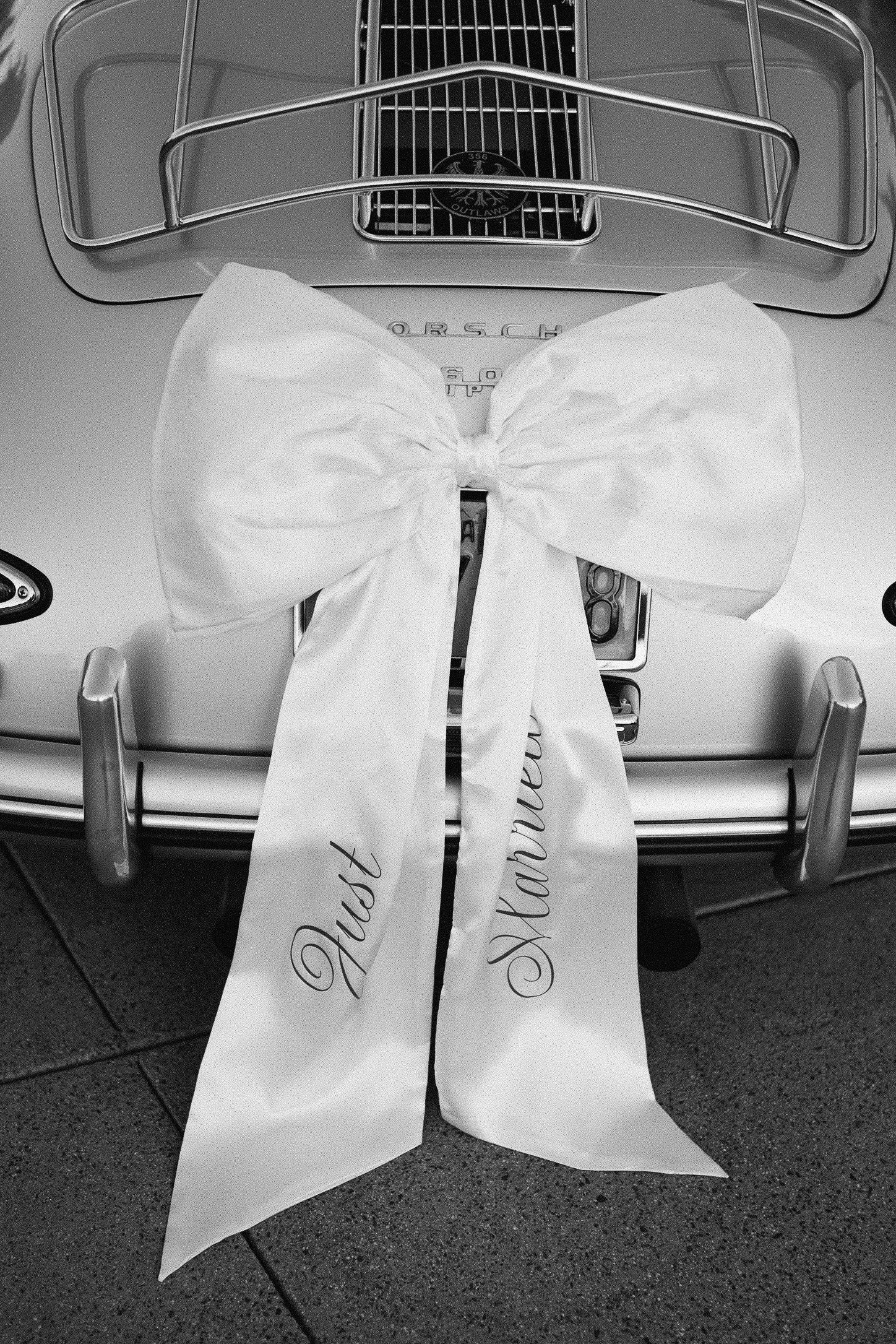 Black and white photo of a classic car adorned with a white satin Just Married bow on the trunk.
