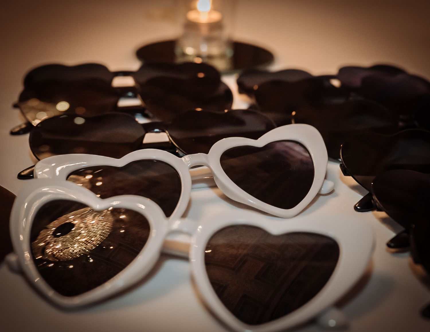 Heart-shaped sunglasses with white and black frames arranged on a table, with a candle glowing softly in the background.