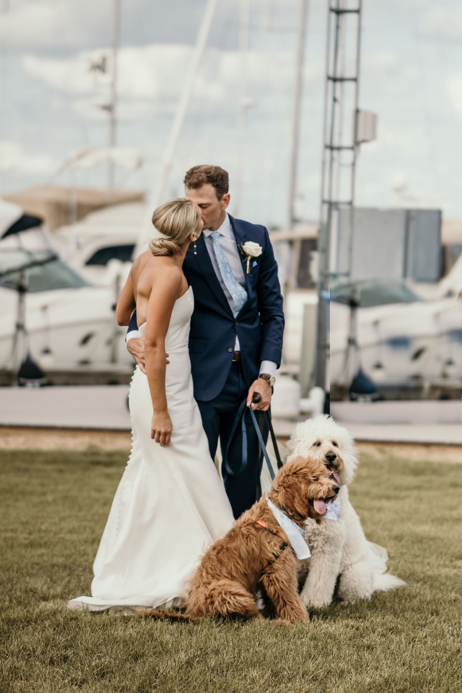 Bride and groom kissing at marina with two goldendoodles wearing bow ties on wedding day.
