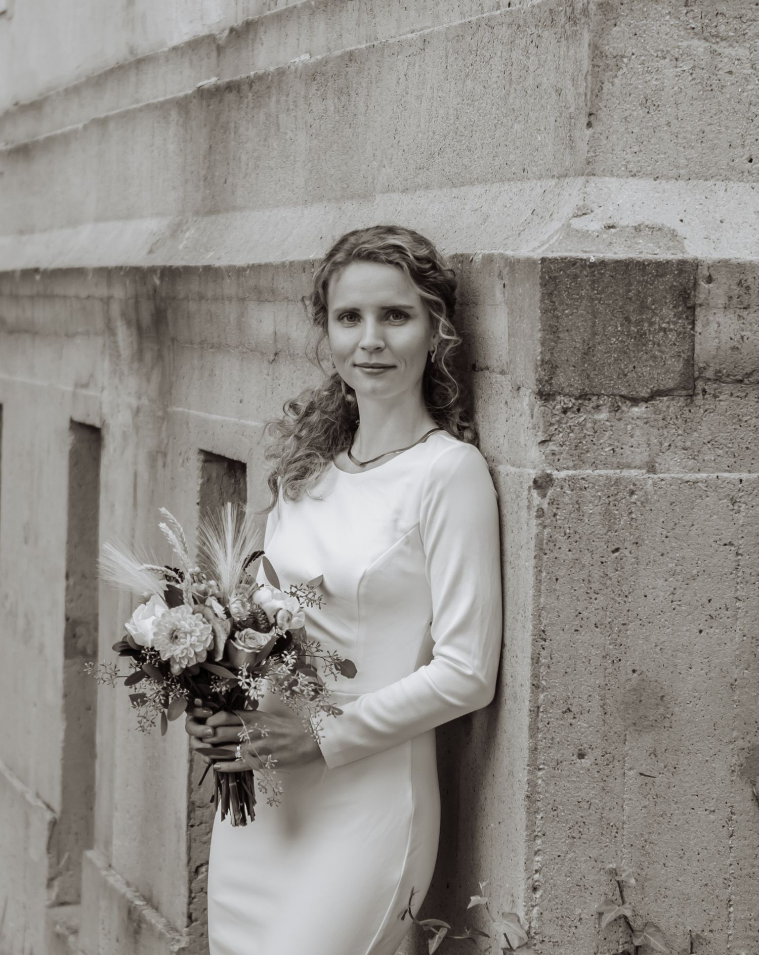 Bride in white long-sleeve gown holding floral bouquet, leaning against stone wall in black and white photo.
