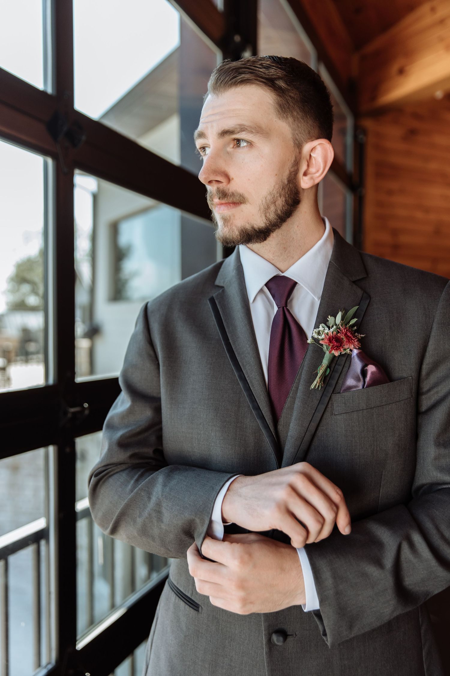 Groom in gray suit with burgundy tie and floral boutonniere adjusting cufflinks by large windows.