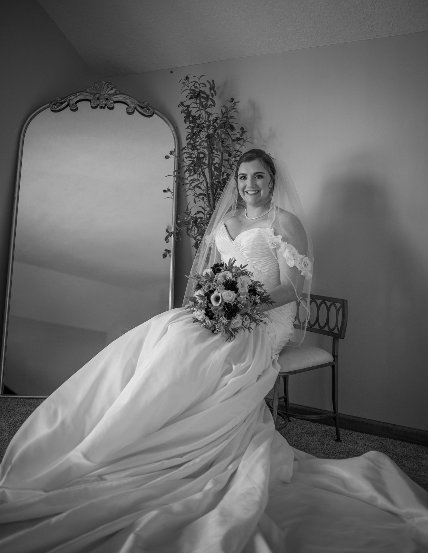 Black and white photo of a smiling bride in a white gown and veil, holding a bouquet, seated near a tall ornate mirror.