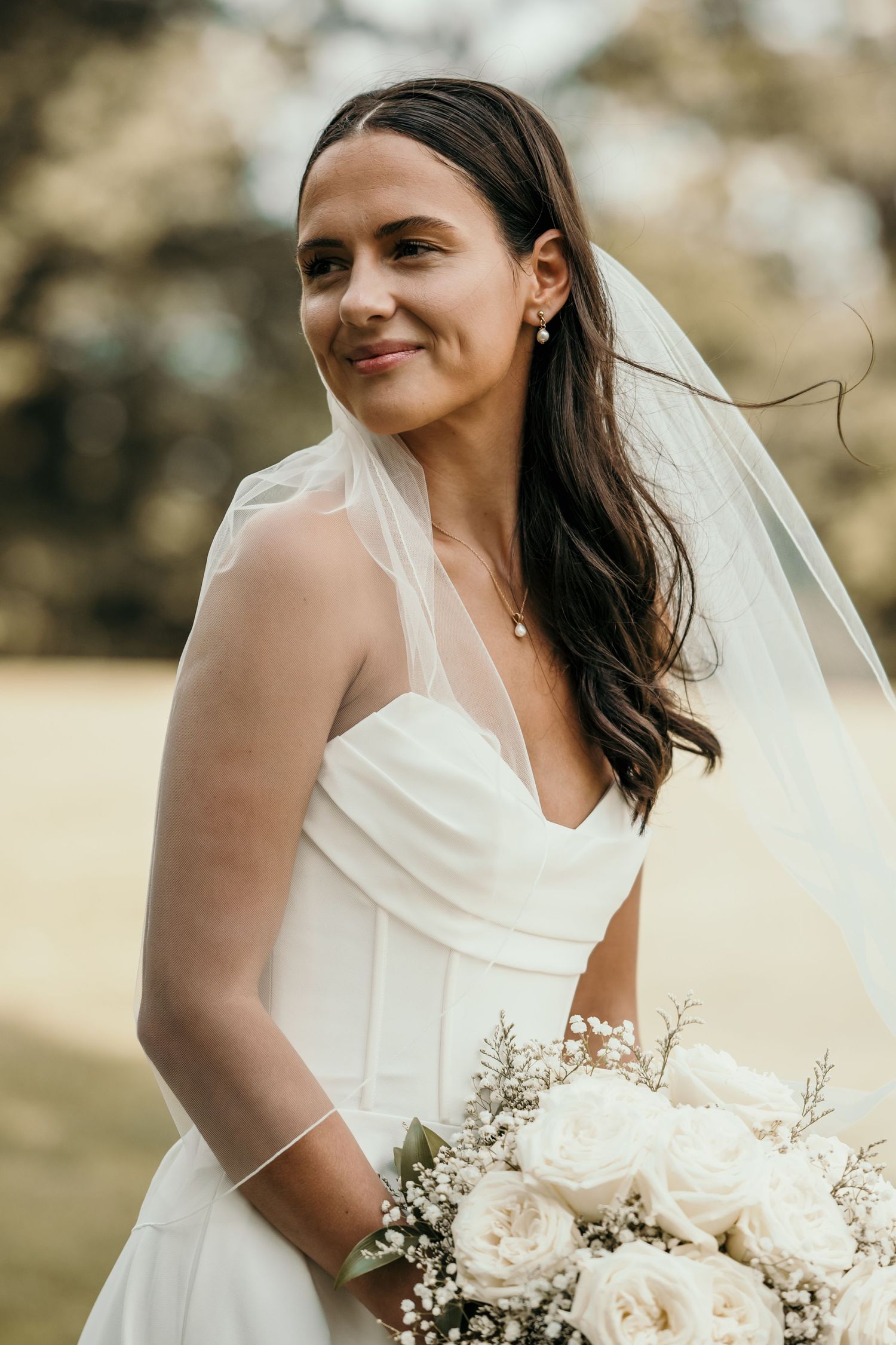 Bride in white gown and veil holds white rose bouquet, smiling outdoors on her wedding day.