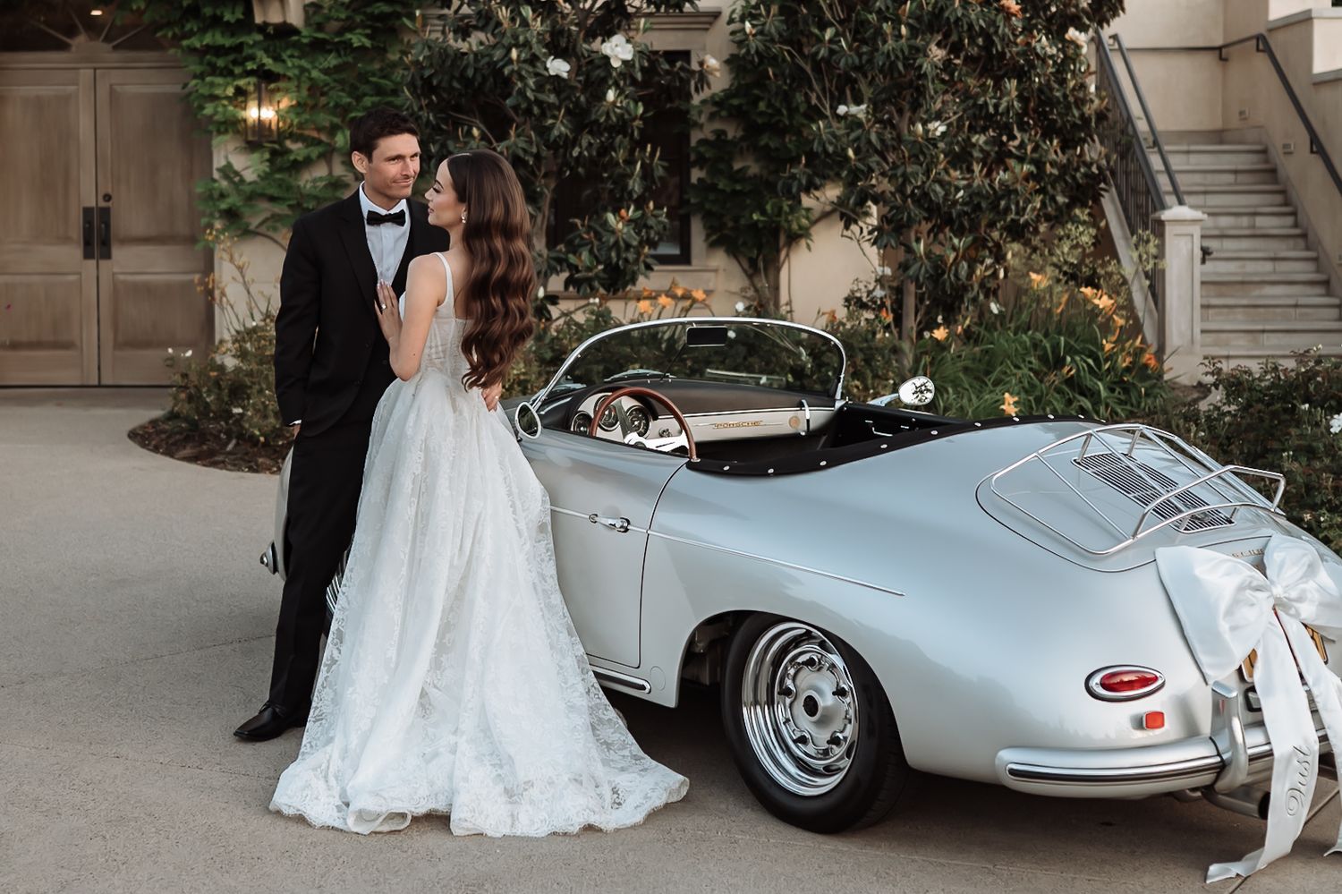 Bride in white gown and groom in tuxedo pose beside a vintage silver Porsche 356 Speedster at elegant wedding venue.