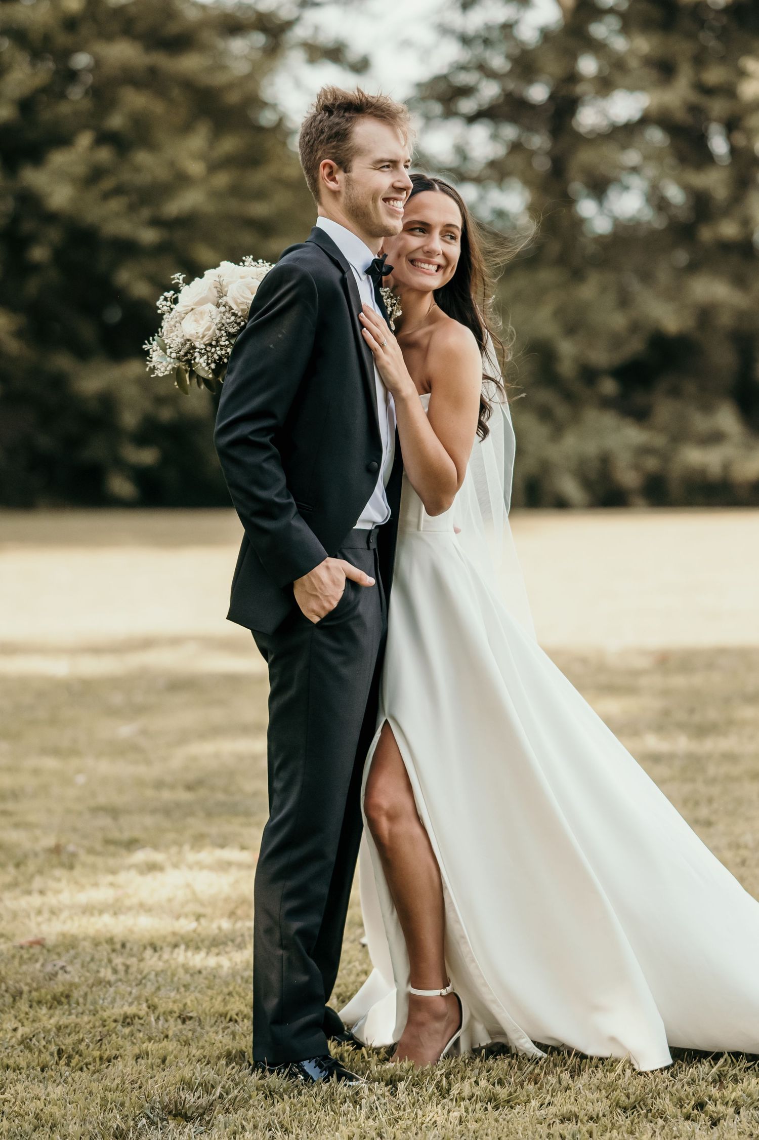 Happy bride in elegant white slit gown embracing groom in navy suit with bouquet in lush green outdoor setting.