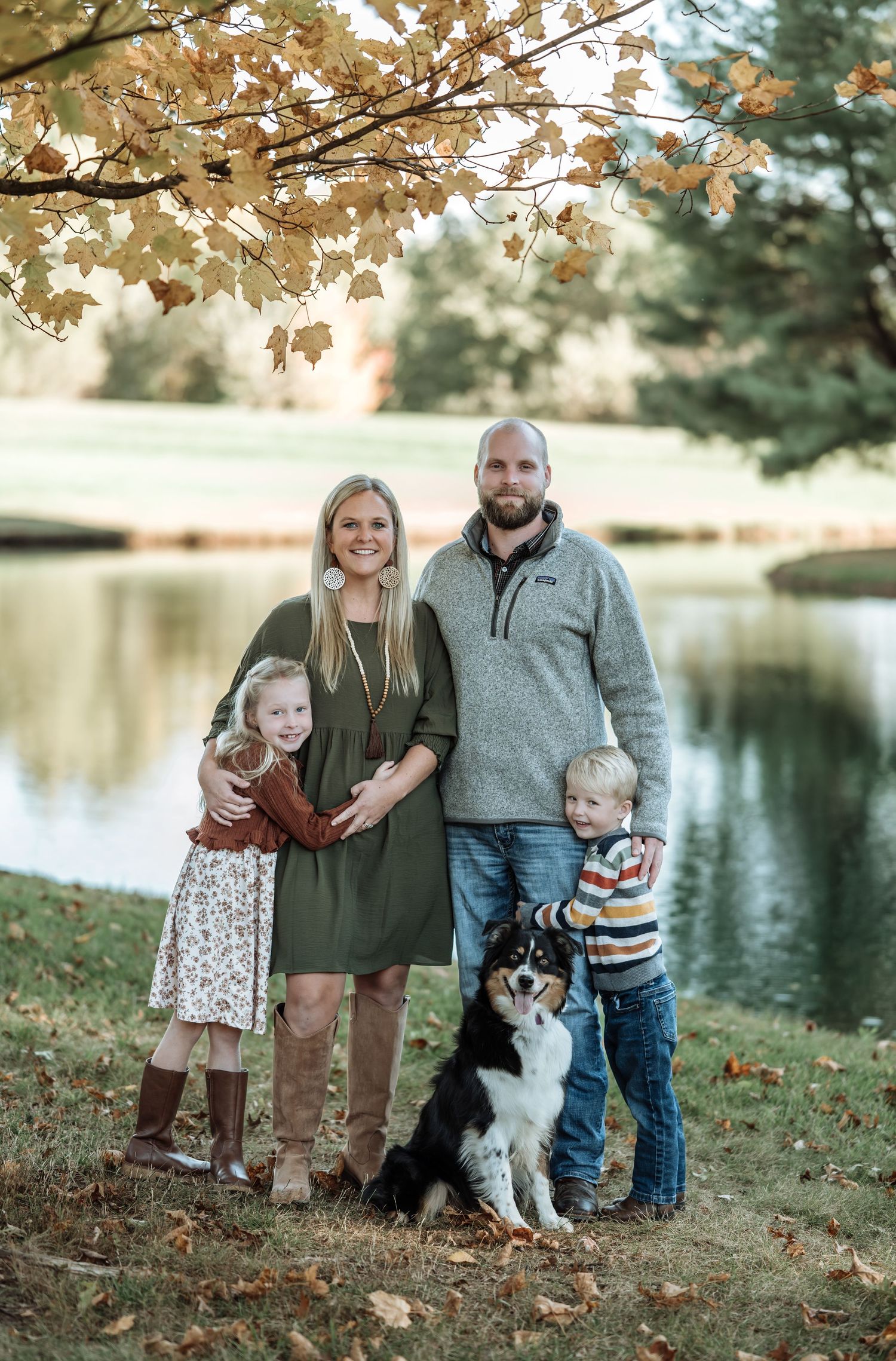 Family of four with a dog posing by a pond under fall foliage.