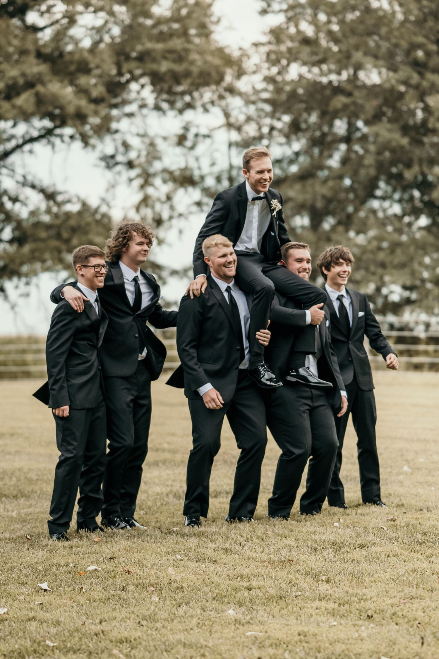 Groomsmen in black suits laughing and lifting groom on shoulders in fun outdoor wedding party photo on grass field.