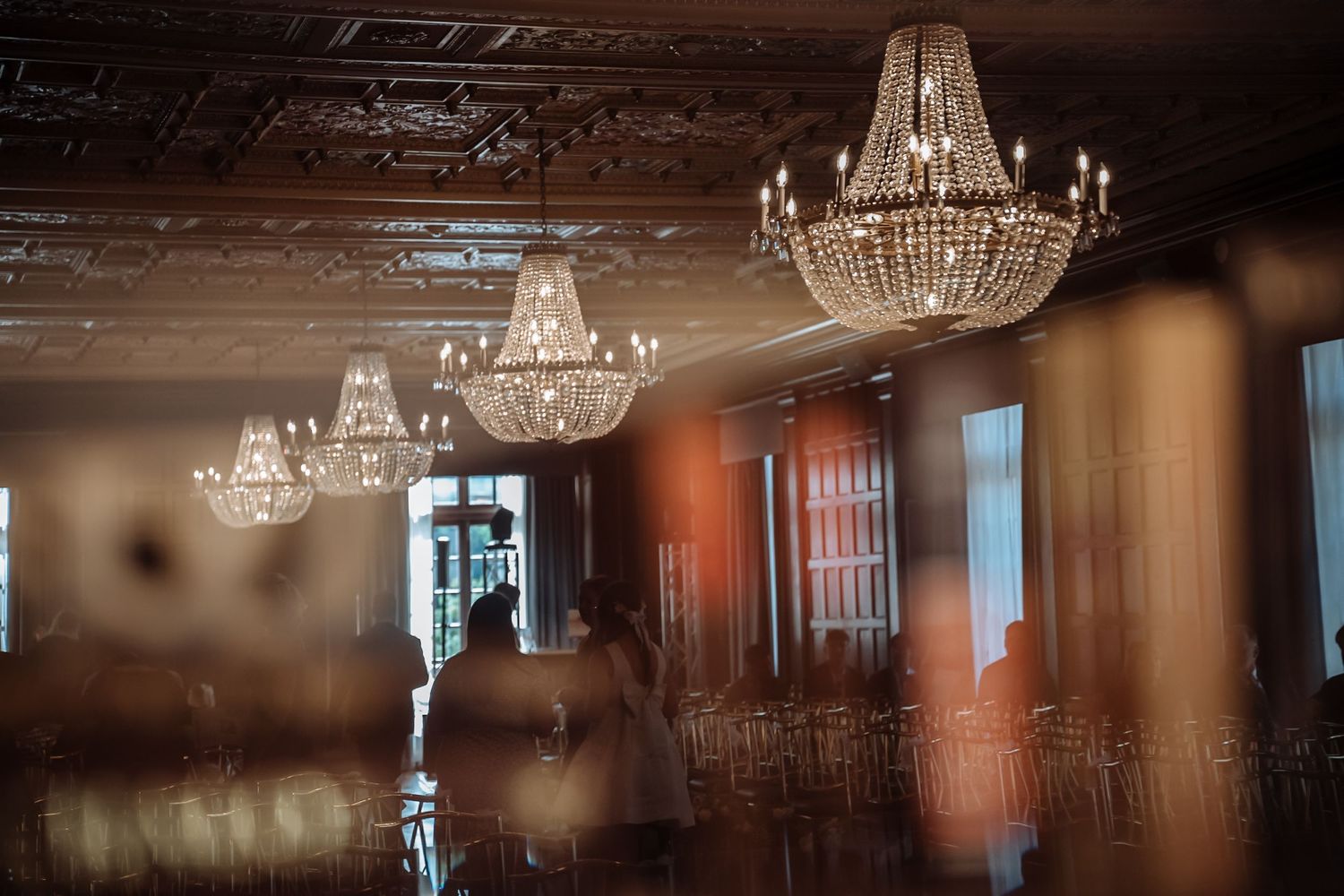 Elegant crystal chandeliers hang from ornate ceiling in luxurious ballroom with warm lighting and reflective surfaces.