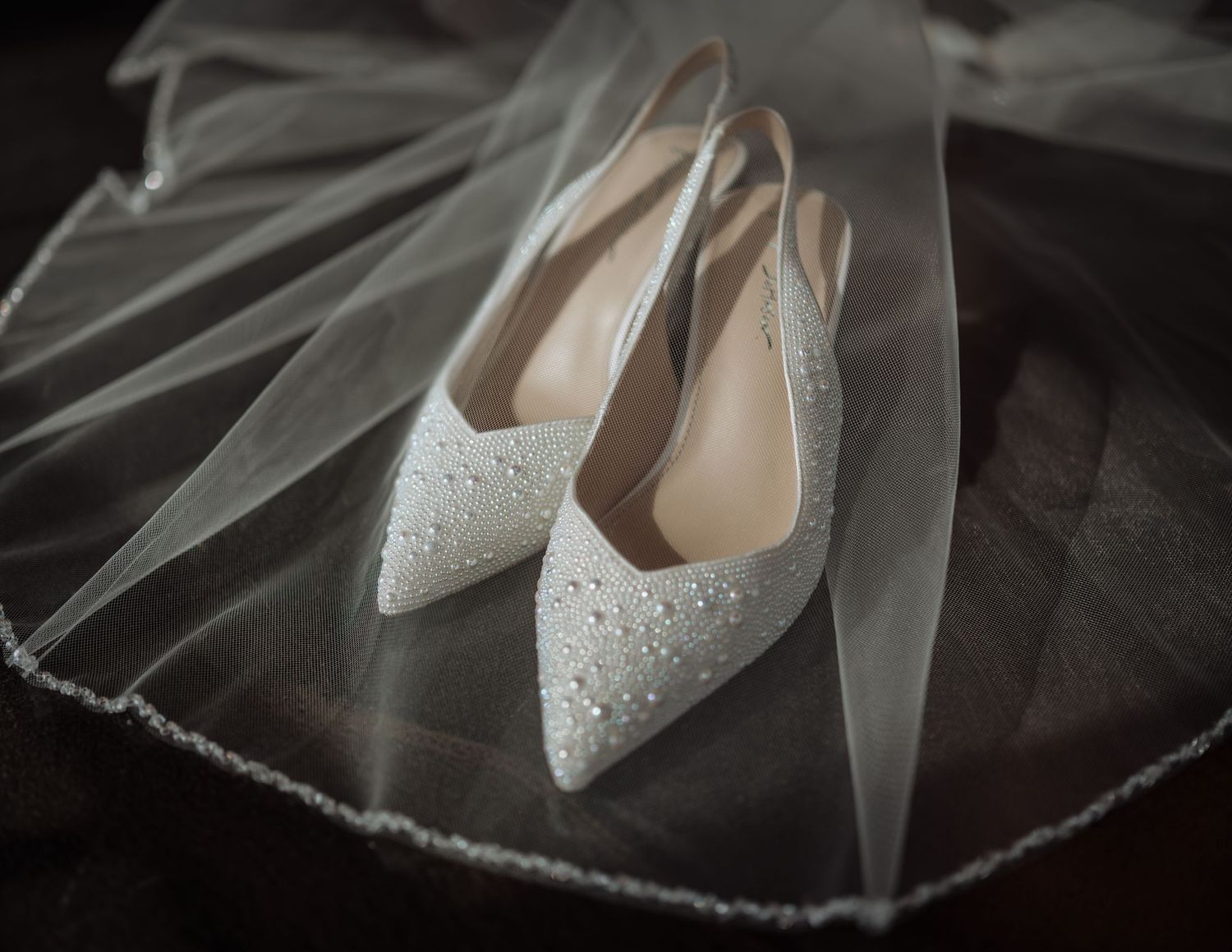 Elegant white beaded bridal heels resting on a sheer wedding veil with pearl trim.