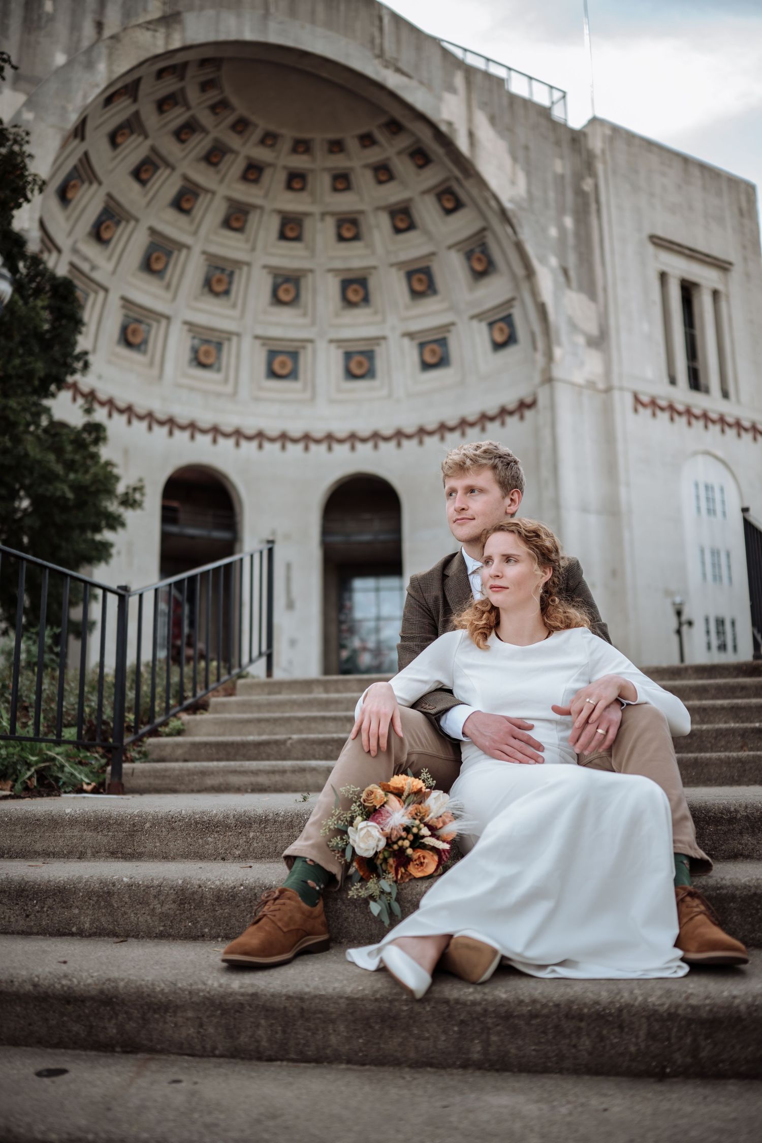 Bride and groom sitting on steps of ornate domed building, bride in white dress holding autumn bouquet.