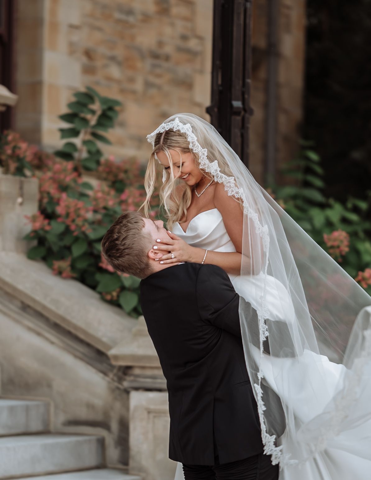 Bride in white gown and lace veil embraced by groom in black tuxedo on stone steps at elegant wedding venue.