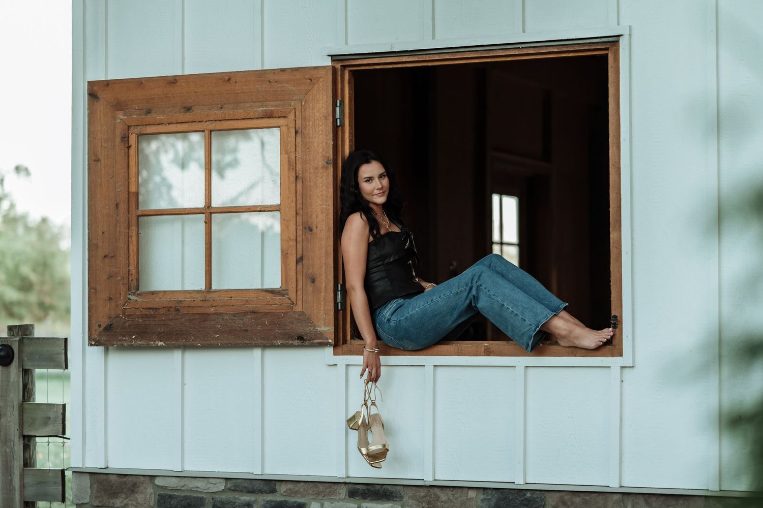 Brunette woman relaxing in wooden barn window frame wearing jeans and black top holding heels.