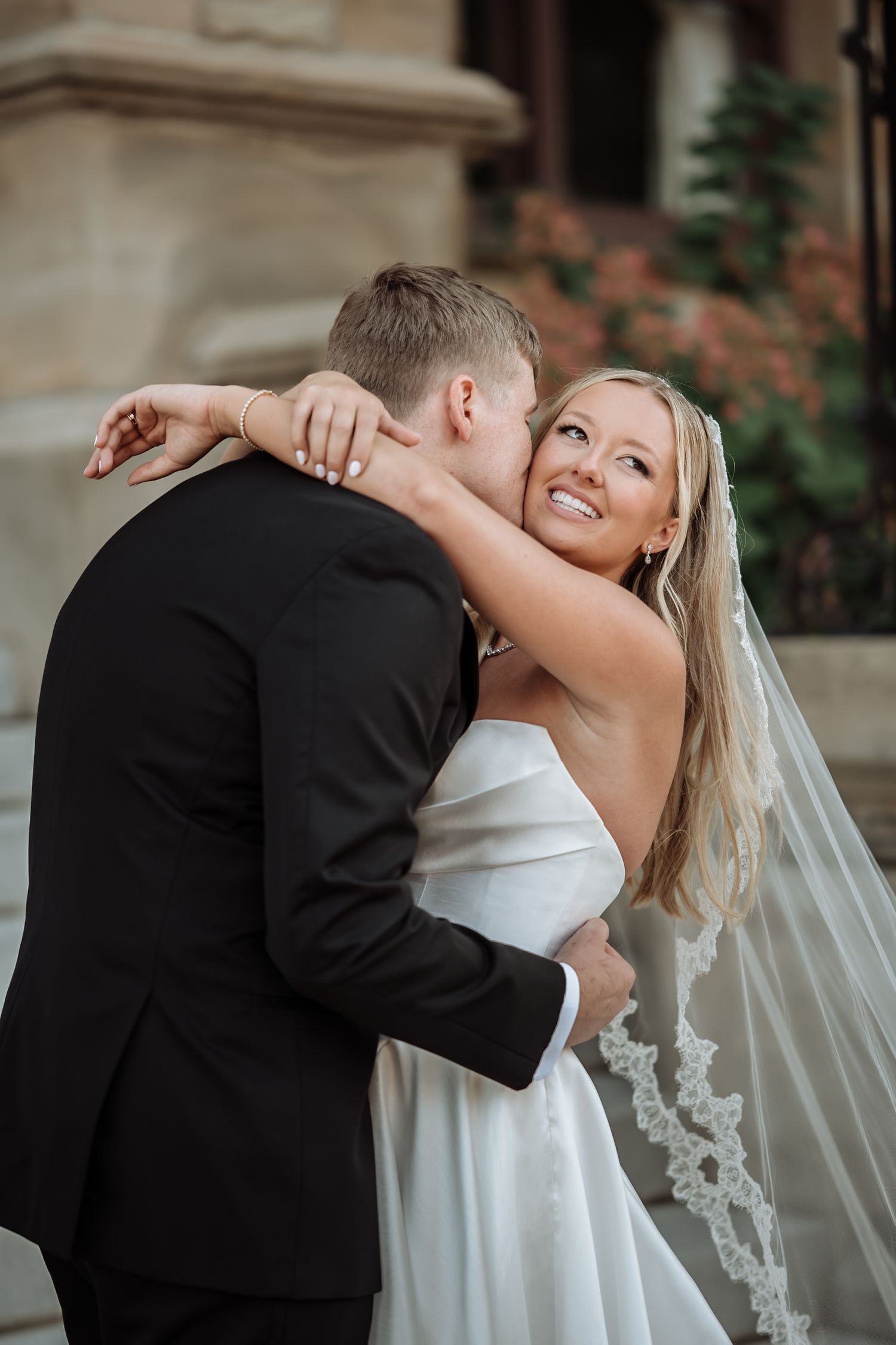A bride in a white gown and veil smiles joyfully as the groom in a black suit kisses her cheek outdoors.