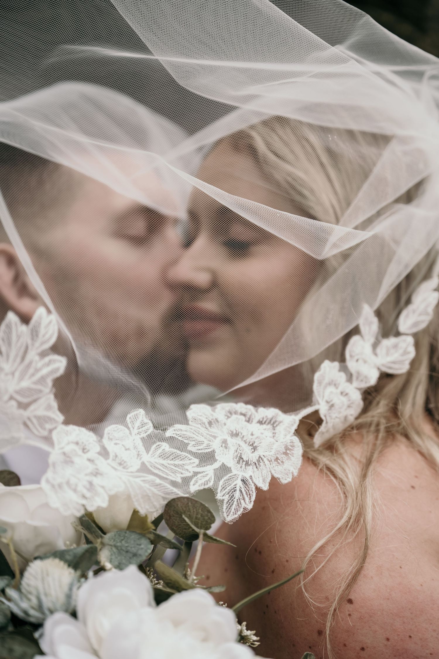 Bride and groom sharing a tender kiss under a lace-trimmed wedding veil, with a white floral bouquet in the foreground.