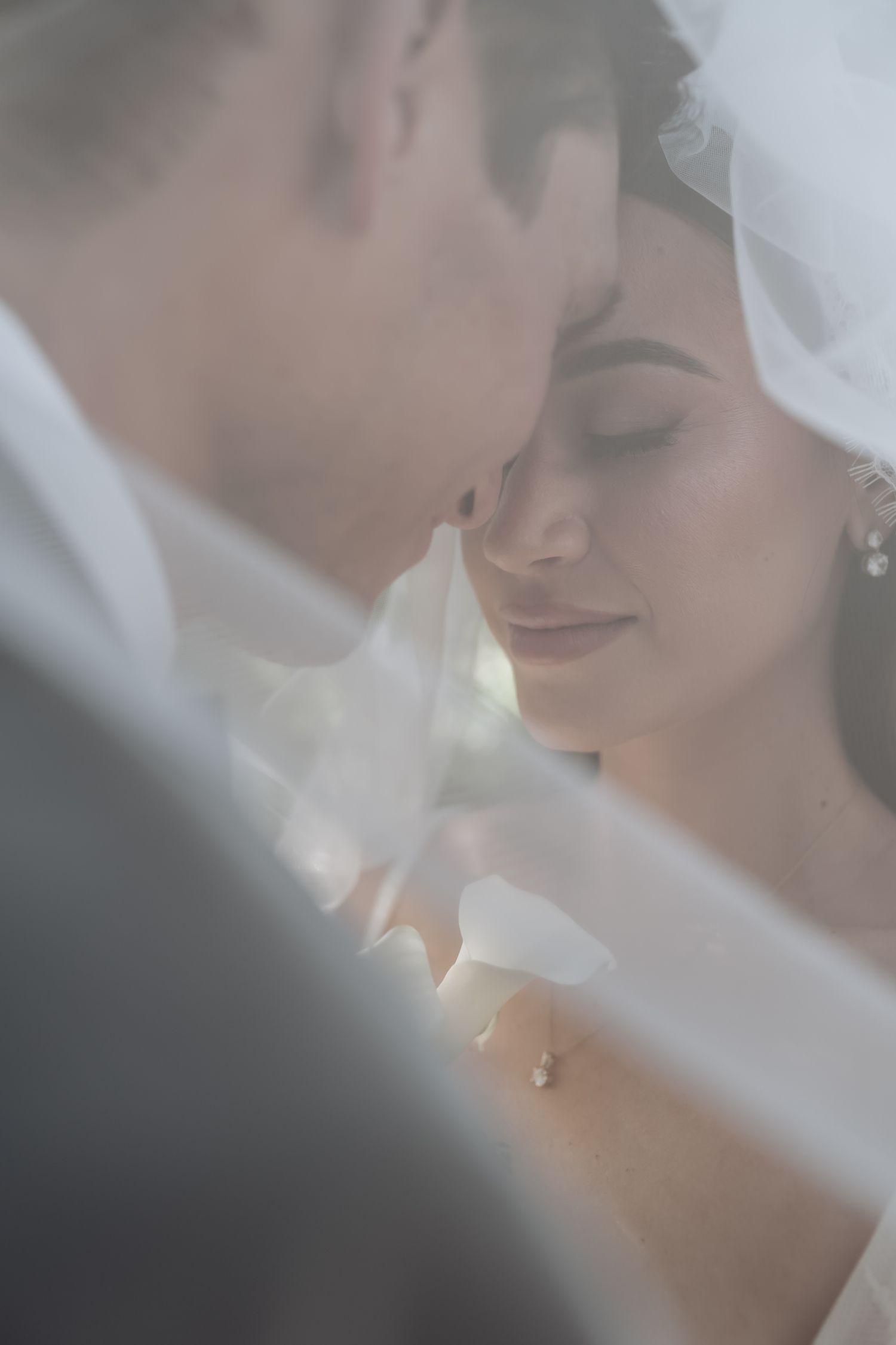 Bride and groom share an intimate moment under a flowing white veil in a soft, dreamy wedding portrait.