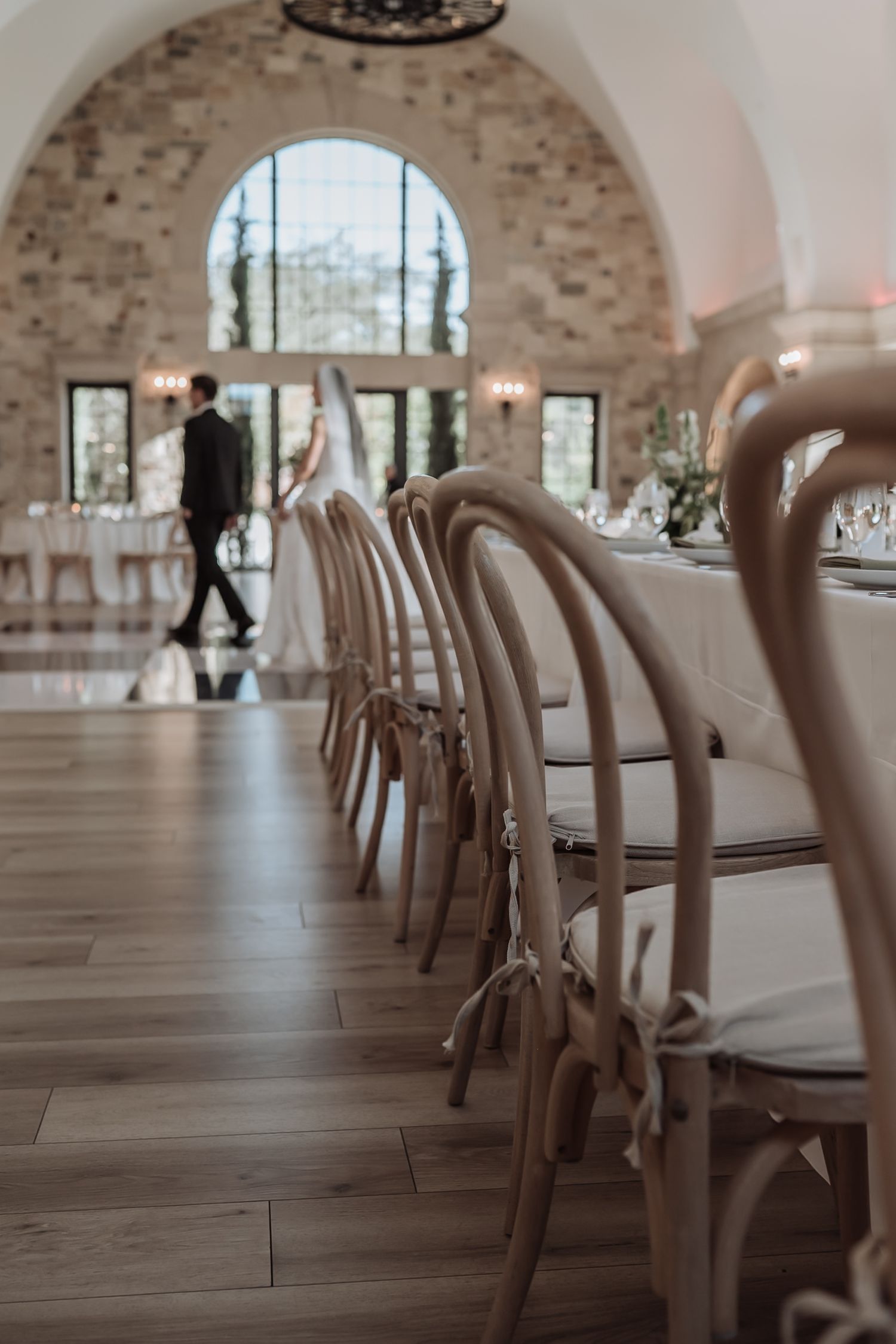 Elegant wedding reception hall with wooden crossback chairs and couple walking in background near arched window.
