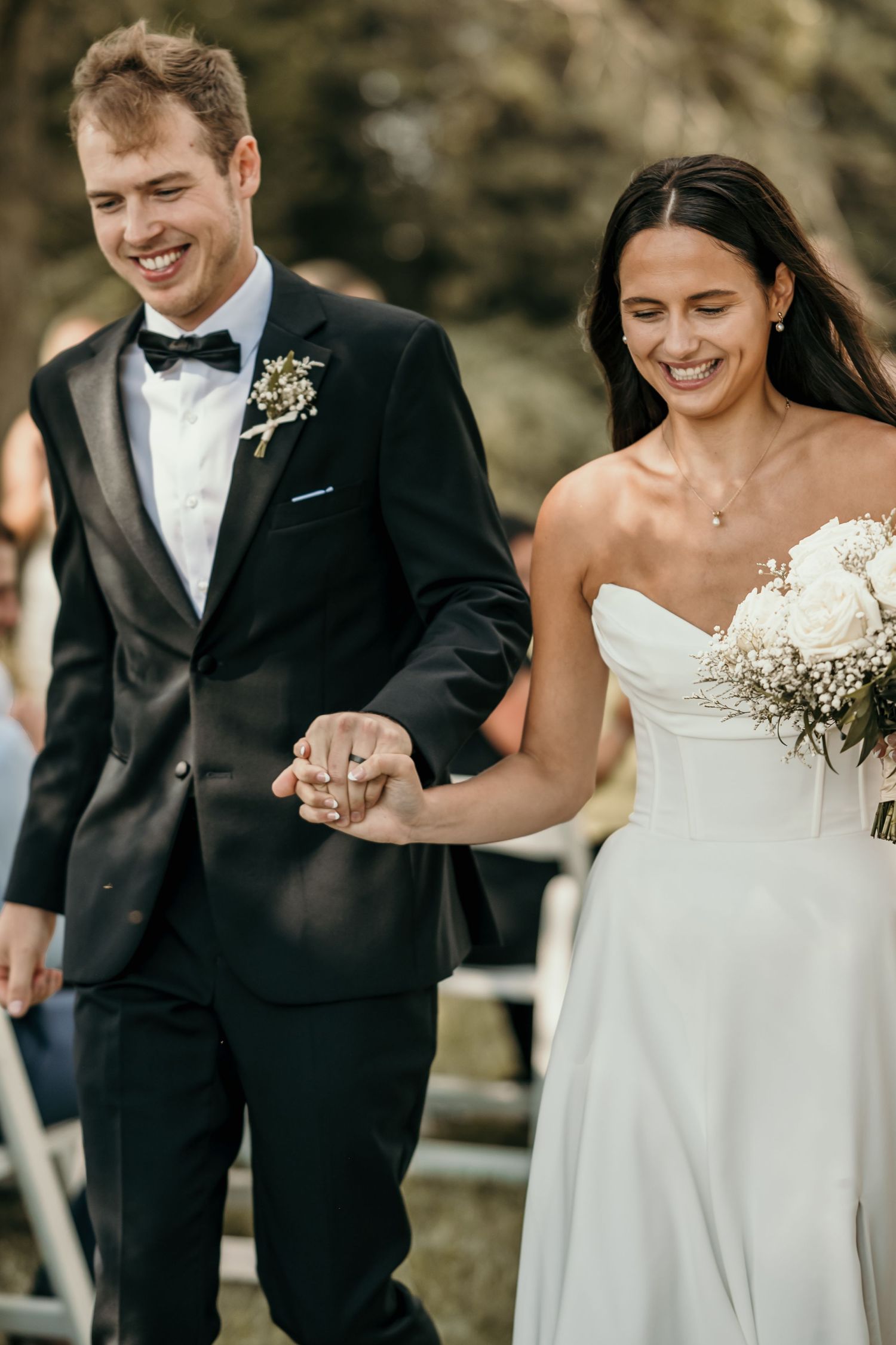 Smiling bride in strapless white gown and groom in black tuxedo holding hands walking down aisle after ceremony.