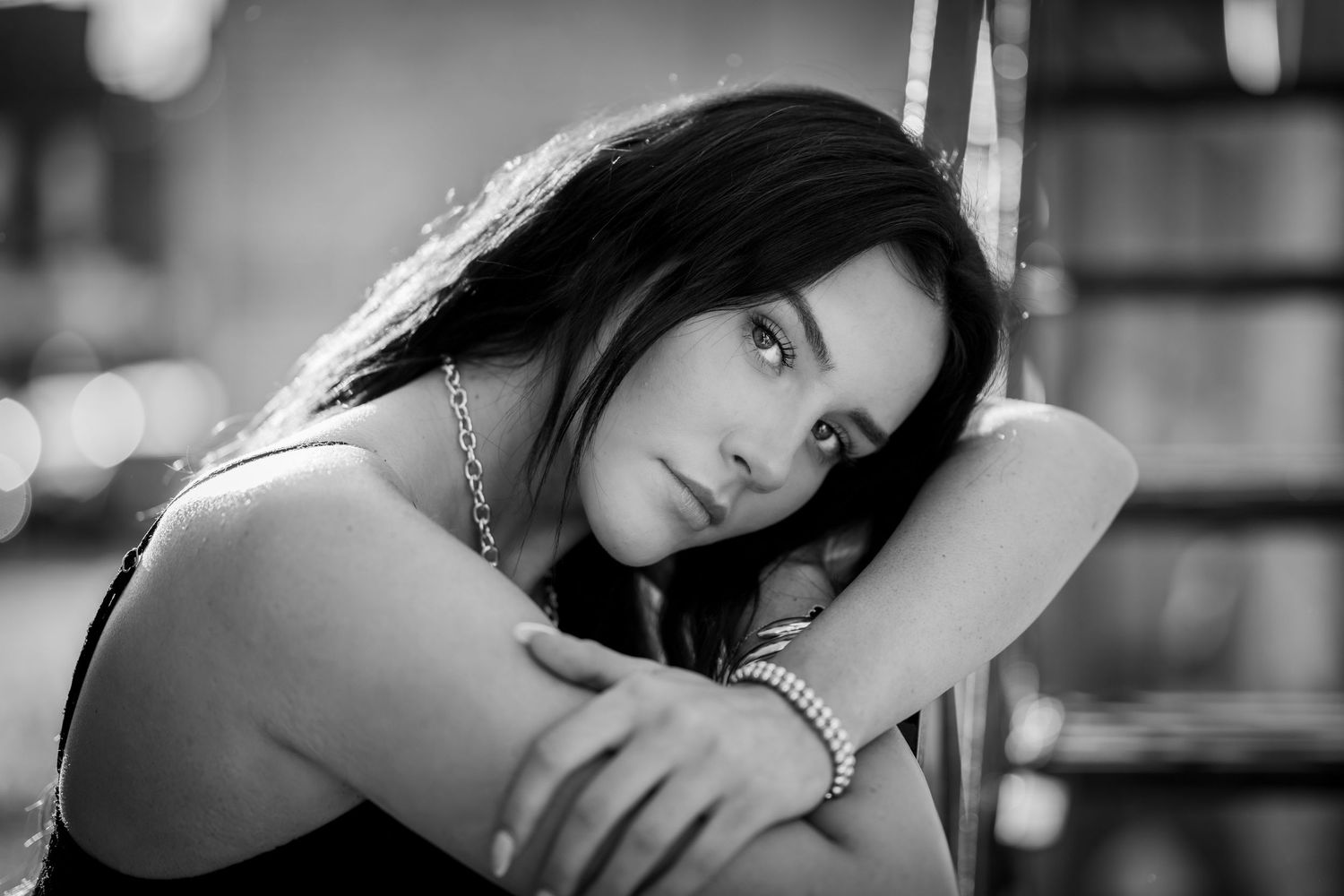 Black and white portrait of brunette woman resting head on arms with bokeh background indoors.