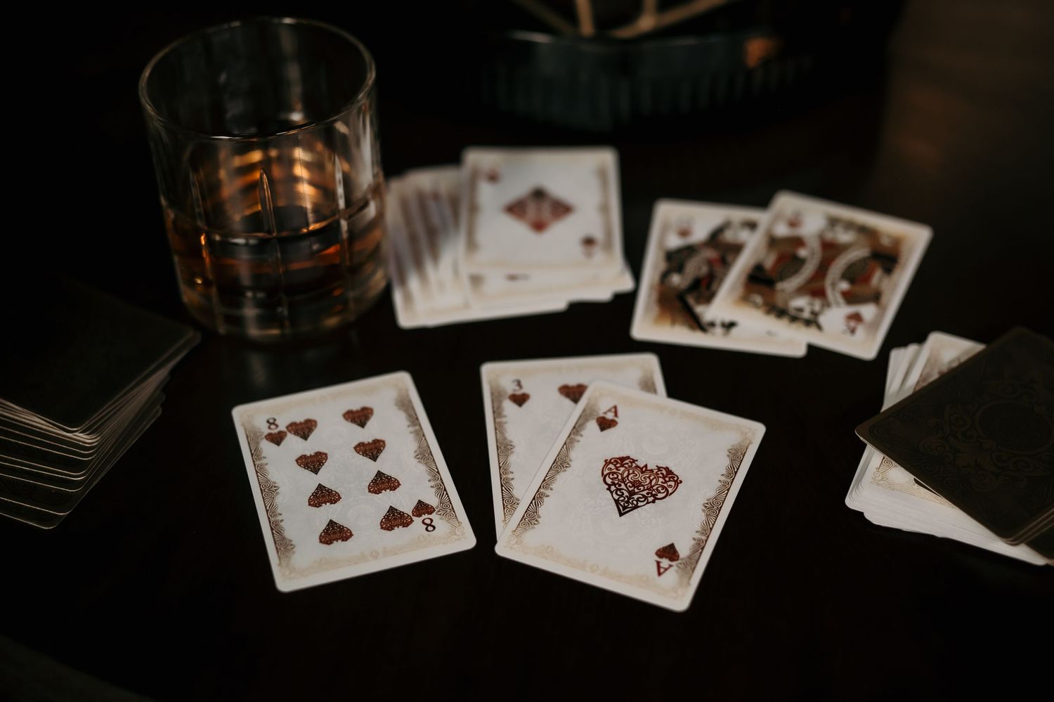 Playing cards spread on a dark table beside a glass of whiskey, featuring queens and tens in an elegant card game setup.