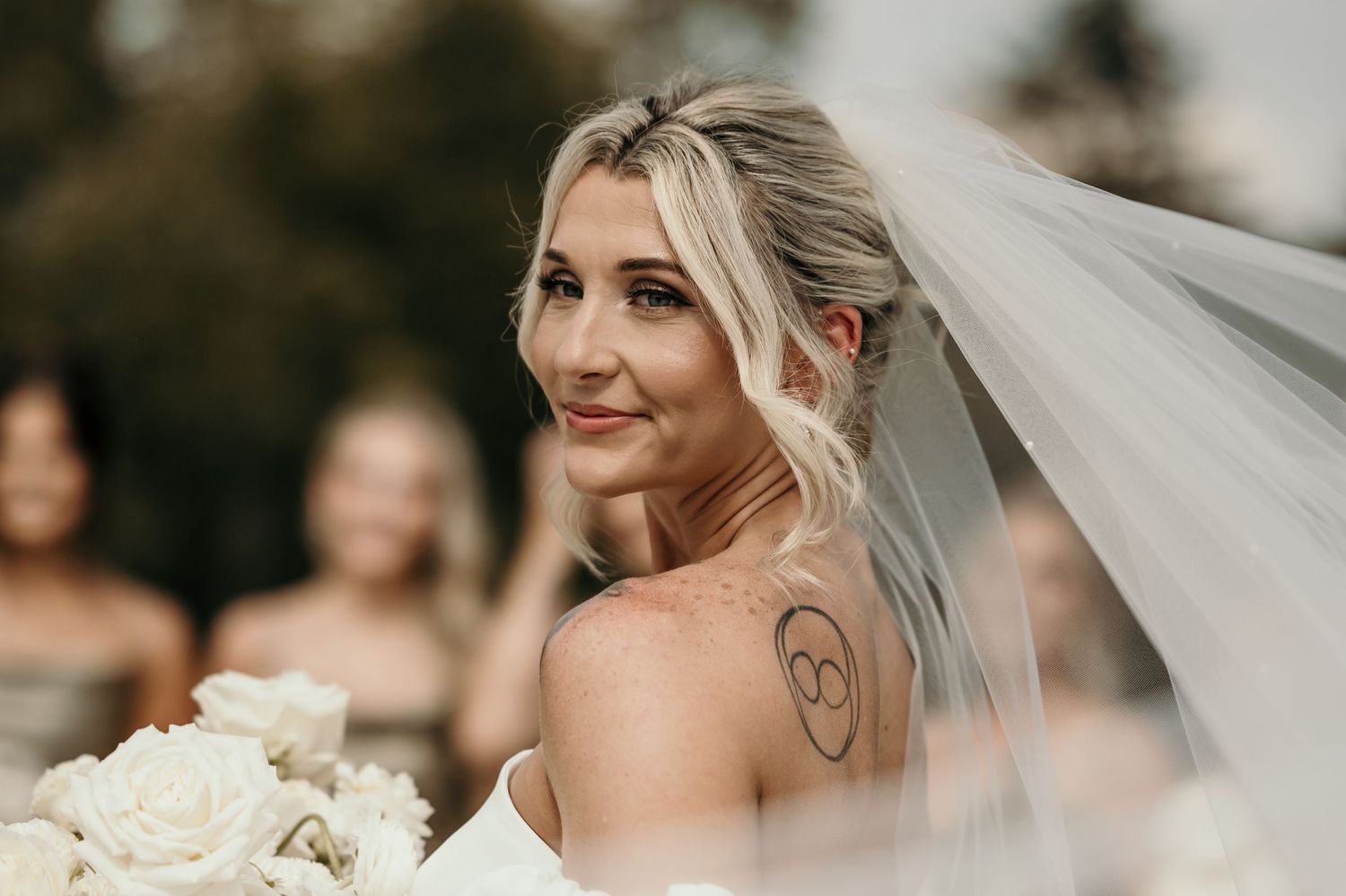 Bride with tattoo smiling over shoulder, wearing veil and holding white bouquet at outdoor wedding ceremony.
