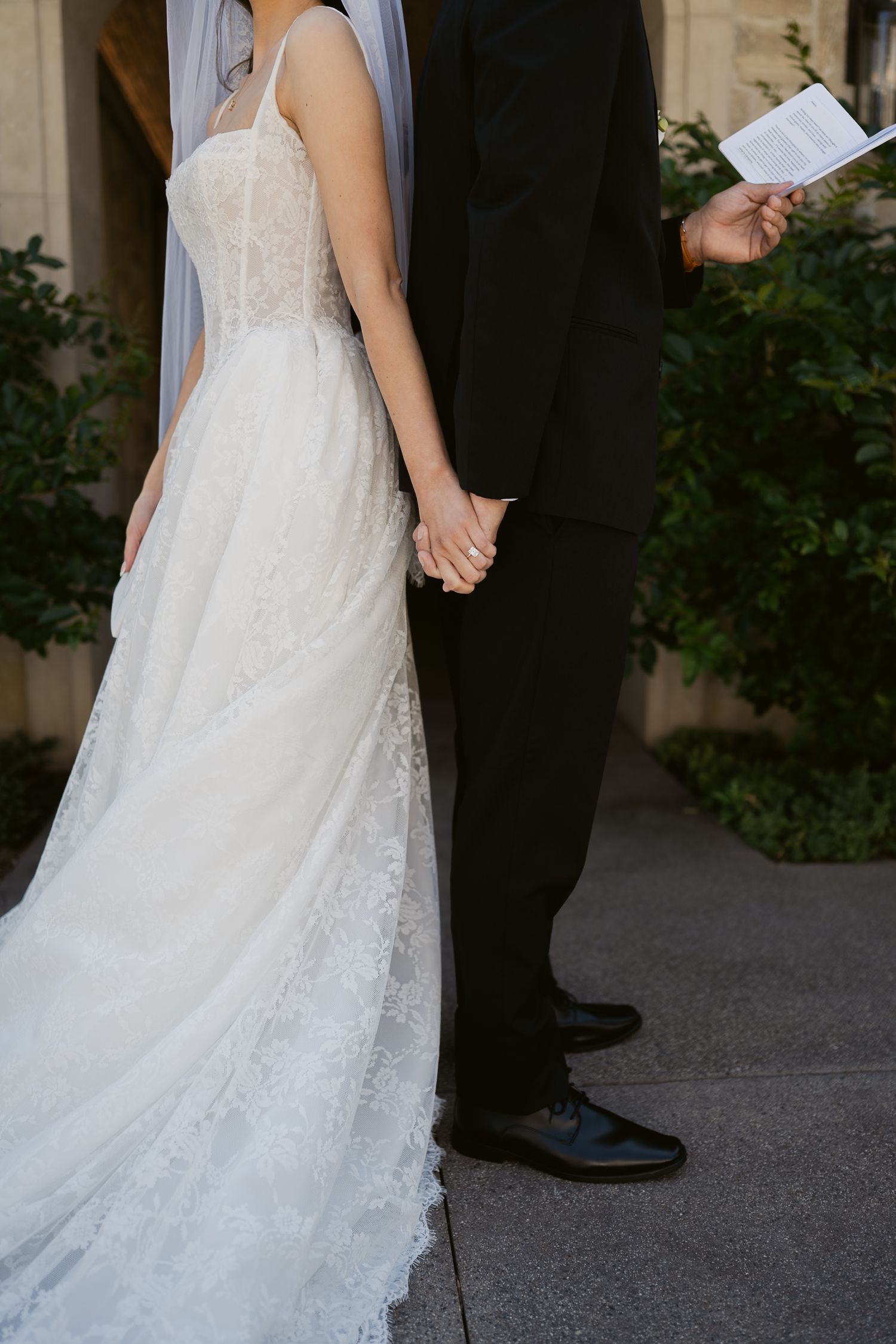 Bride in elegant lace A-line wedding gown holding hands with groom in black suit during outdoor ceremony.