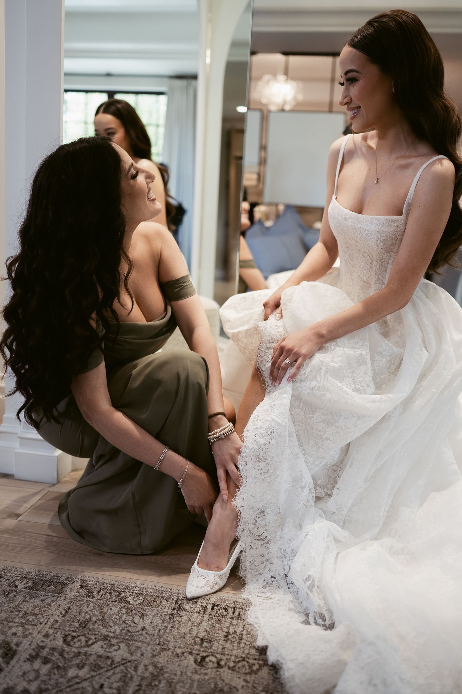 Bridesmaid helping bride put on white heels while wearing lace wedding gown.