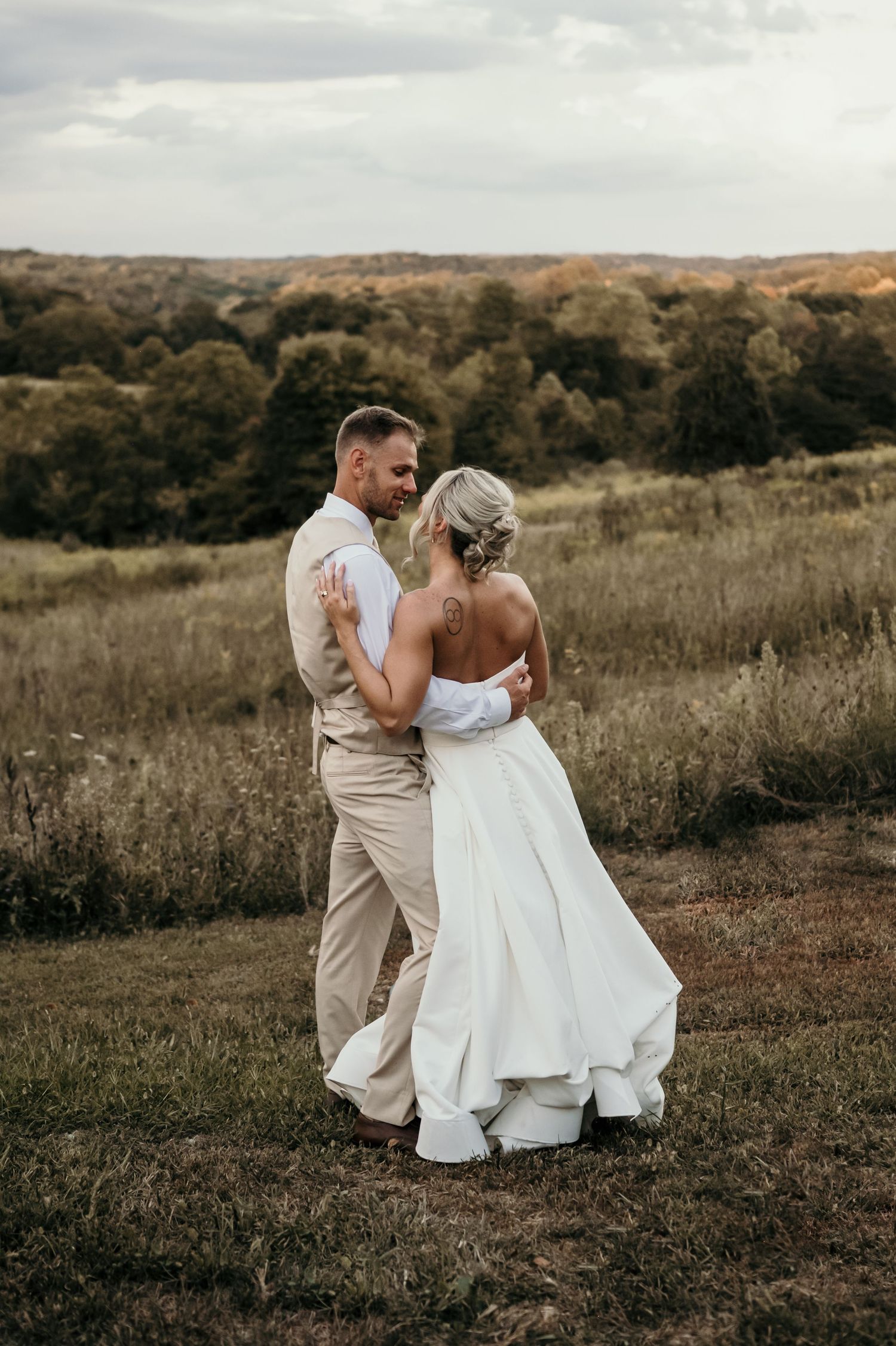 Bride and groom share a romantic dance in a scenic outdoor field with lush green trees and rolling hills at sunset.