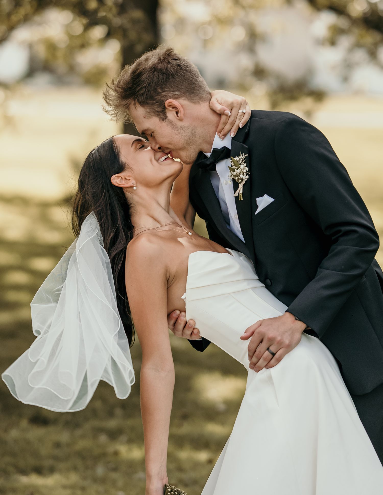 Groom in black tuxedo dips and kisses his bride in a white strapless gown outdoors.
