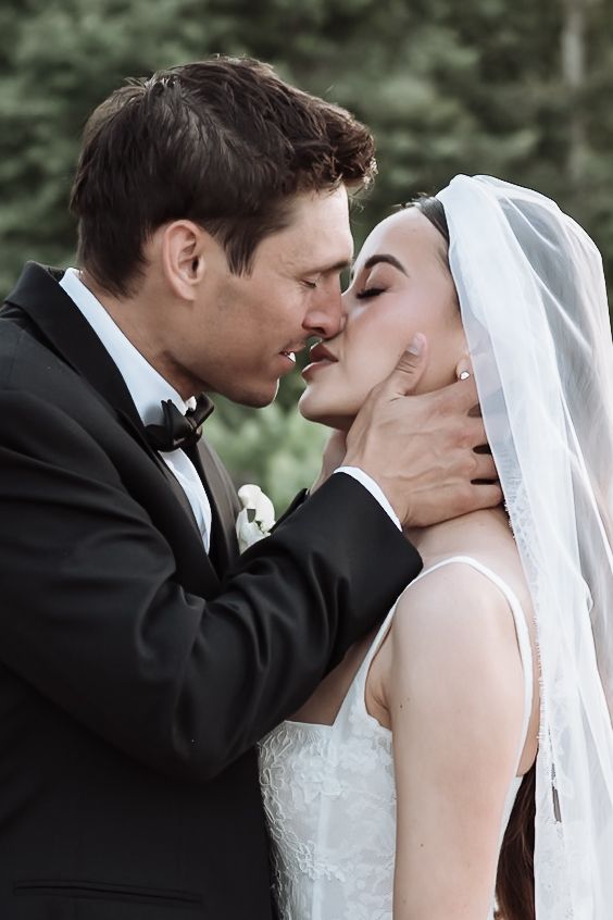 Groom in black tuxedo gently holds brides face as they share a romantic kiss during outdoor wedding ceremony.