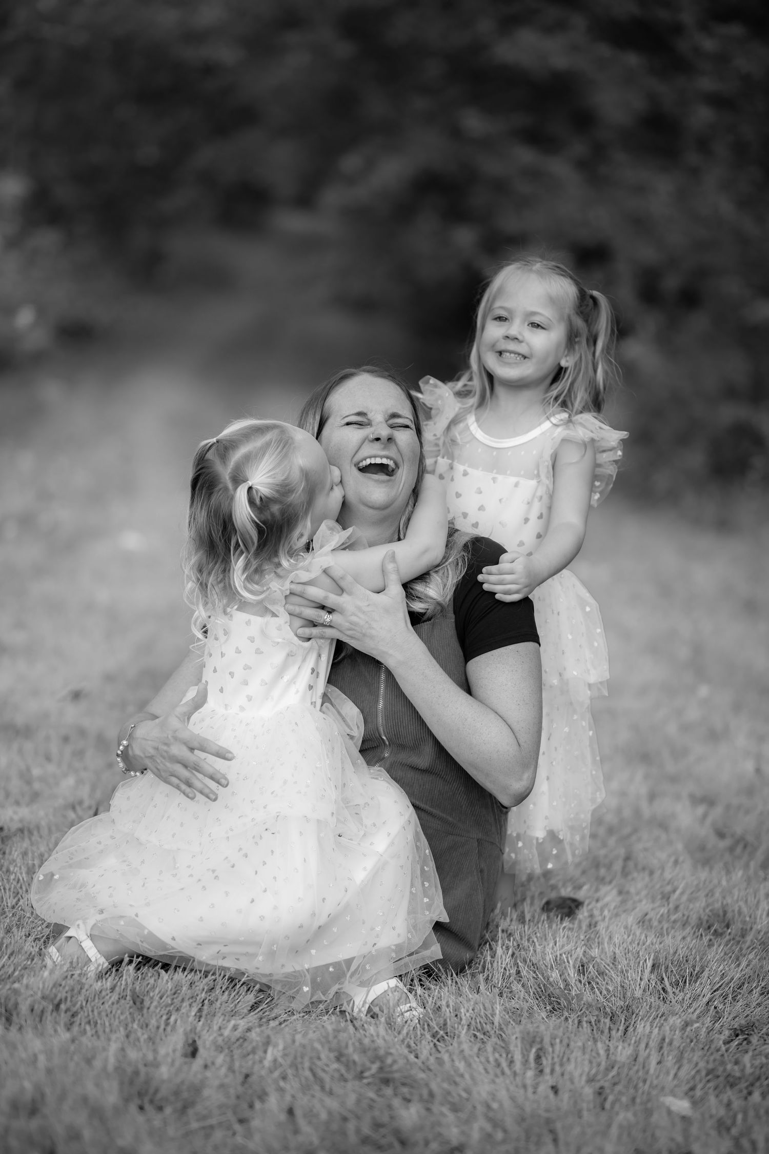 Black and white photo of mother laughing joyfully while hugging two young daughters in white dresses.