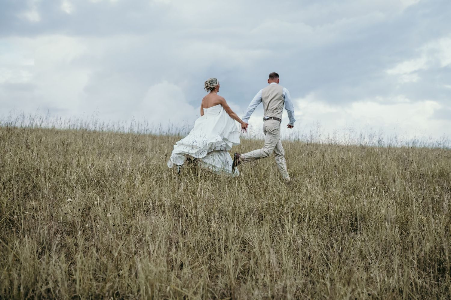 A bride and groom run hand-in-hand through a golden grass field under a cloudy sky on their wedding day.