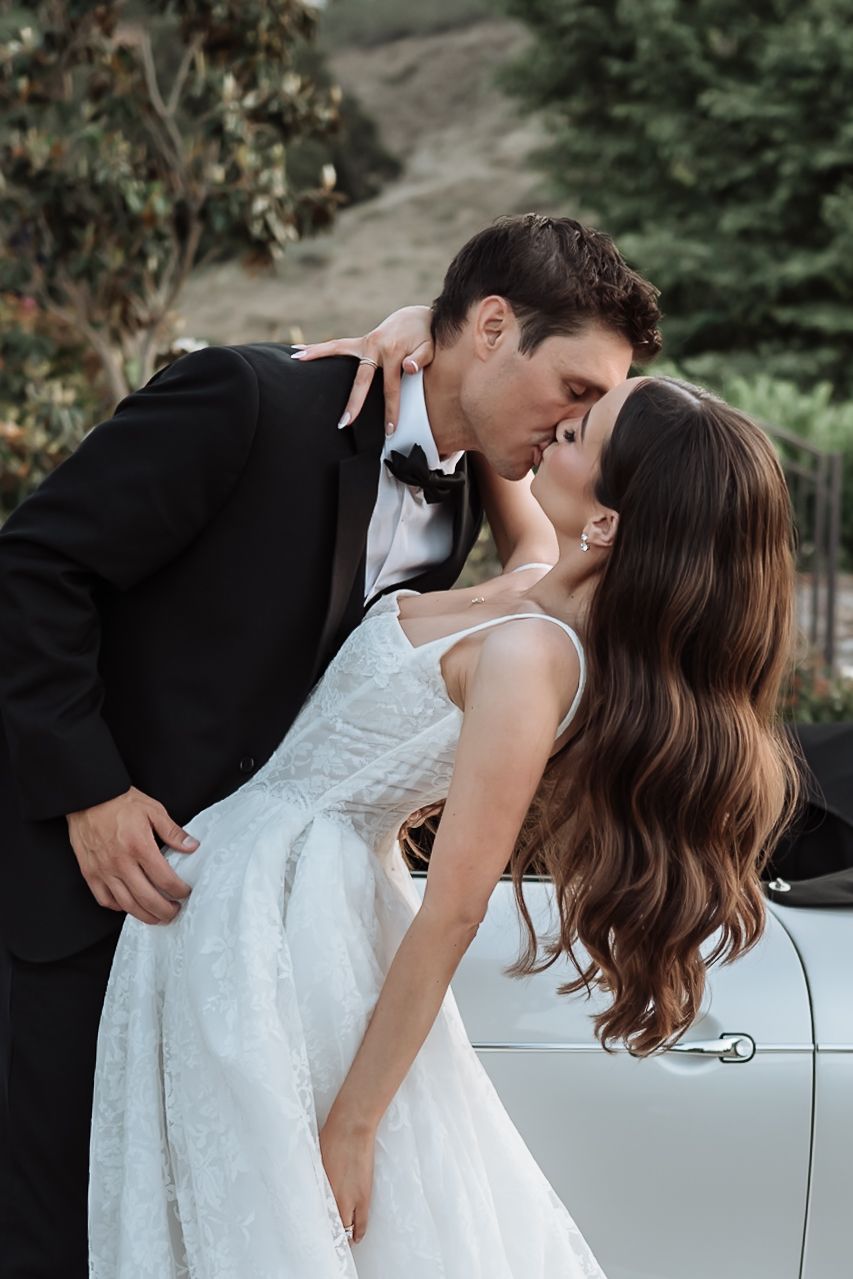Bride and groom sharing romantic kiss beside vintage white car outdoors.