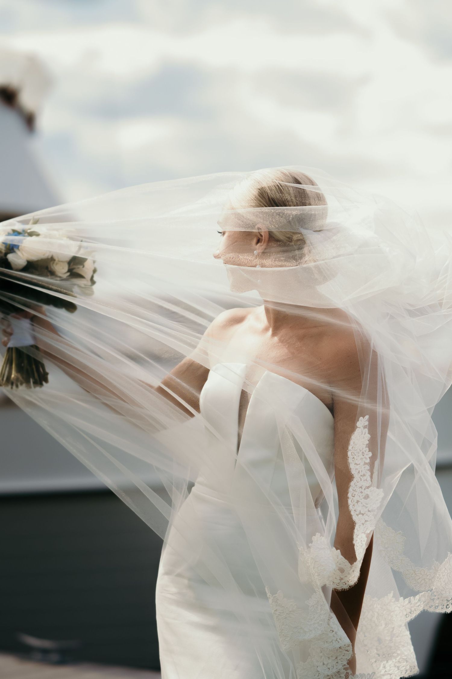 Bride in strapless white gown with flowing veil blowing in wind, holding bouquet against cloudy sky.