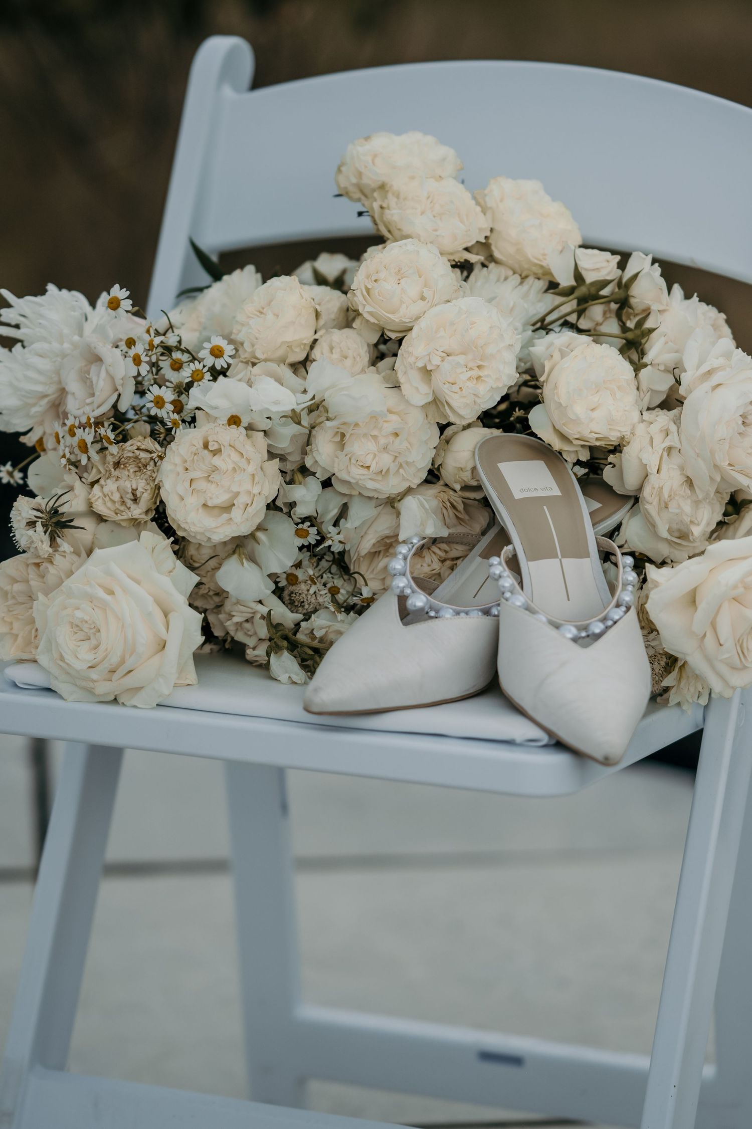White bridal heels with pearl details rest on a white chair beside an elegant bouquet of cream roses and wildflowers.