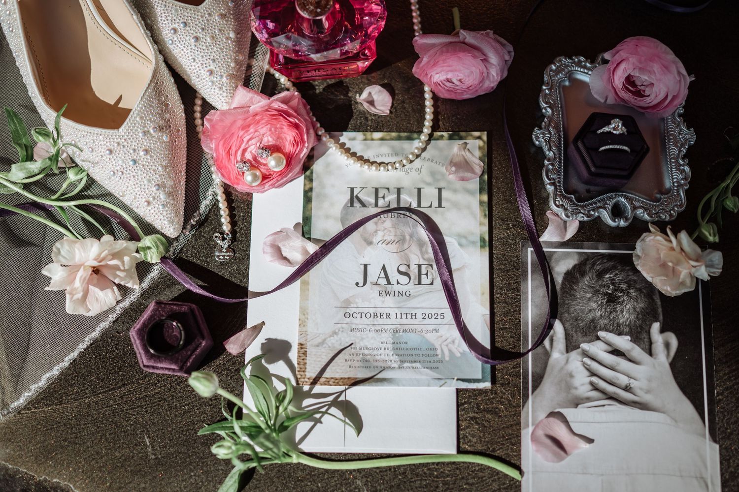 Elegant wedding flat lay featuring floral invitations, pink ranunculus, pearl jewelry, and ring boxes on a reflective surface.