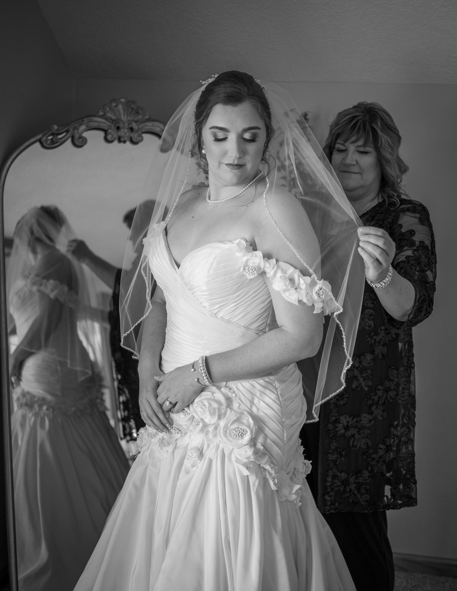 Black and white photo of a bride in a strapless gown and veil being assisted by a woman near an ornate mirror.